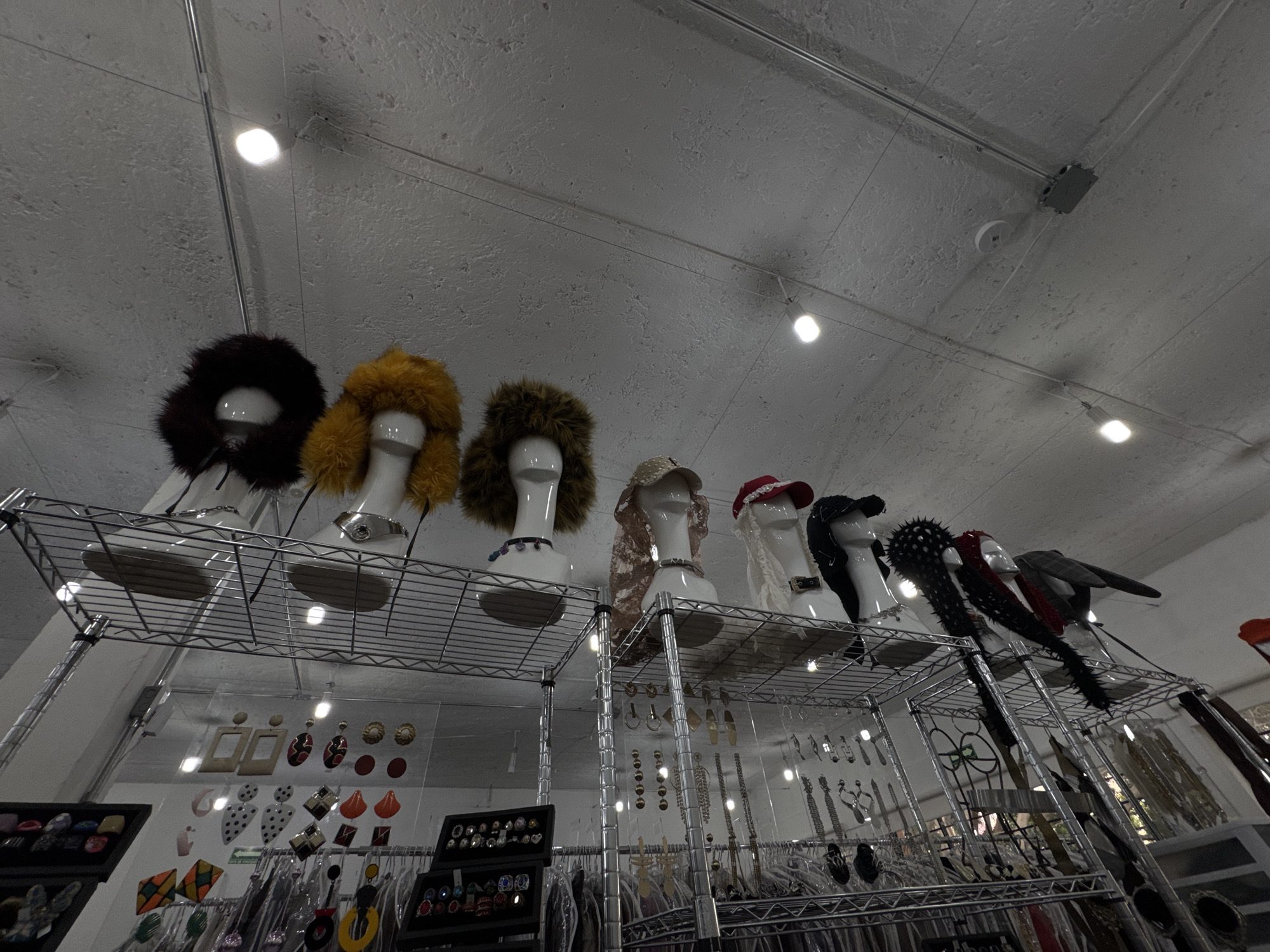 The image captures an indoor retail space, possibly a clothing or accessory store, in Mexico City, Mexico. Seven mannequin heads are displayed on a wire shelving unit. Each mannequin wears a different hat or headpiece, ranging from faux fur hoods to various styles of hats adorned with embellishments like spikes and feathers. Behind the mannequins, a collection of jewelry, accessories, and clothing hang on racks, suggesting a curated selection of fashion items. The ceiling has a textured finish and is illuminated by several recessed lights. The overall atmosphere is slightly muted due to the lighting.