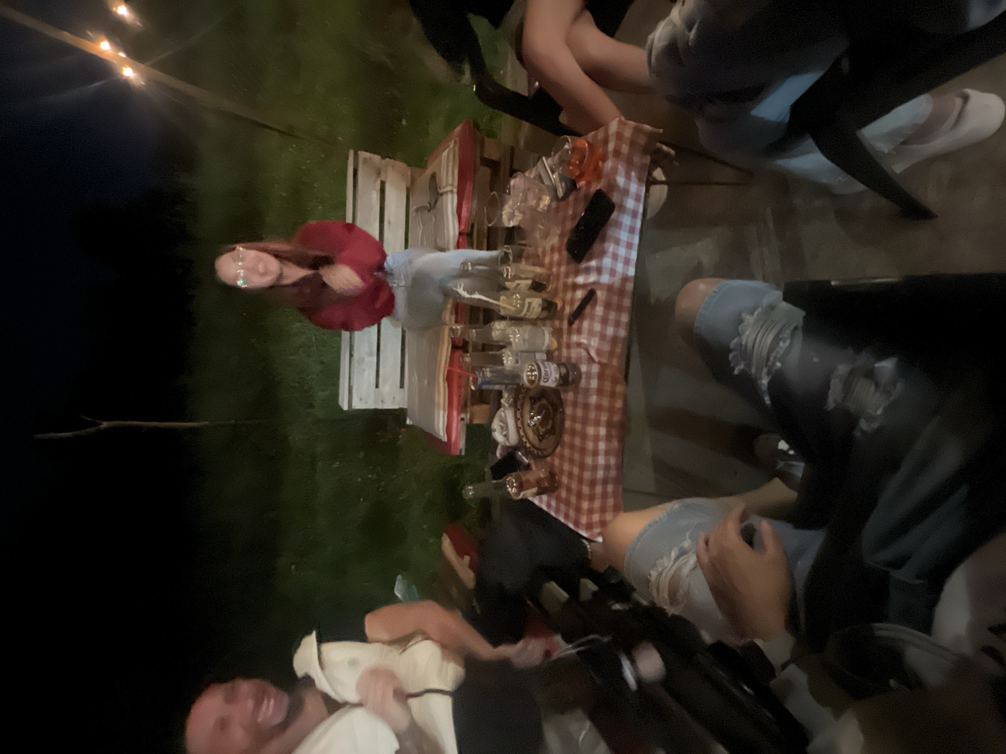 A gathering is occurring outdoors at night. A woman in glasses and a red shirt stands near a table covered with a red-and-white checkered cloth, upon which there are beer bottles and glasses. Several people are seated around the table; one individual is wearing torn jeans. String lights are visible in the background. The setting appears to be grassy. I am unable to determine the location in the city of Касапите, Bulgaria.