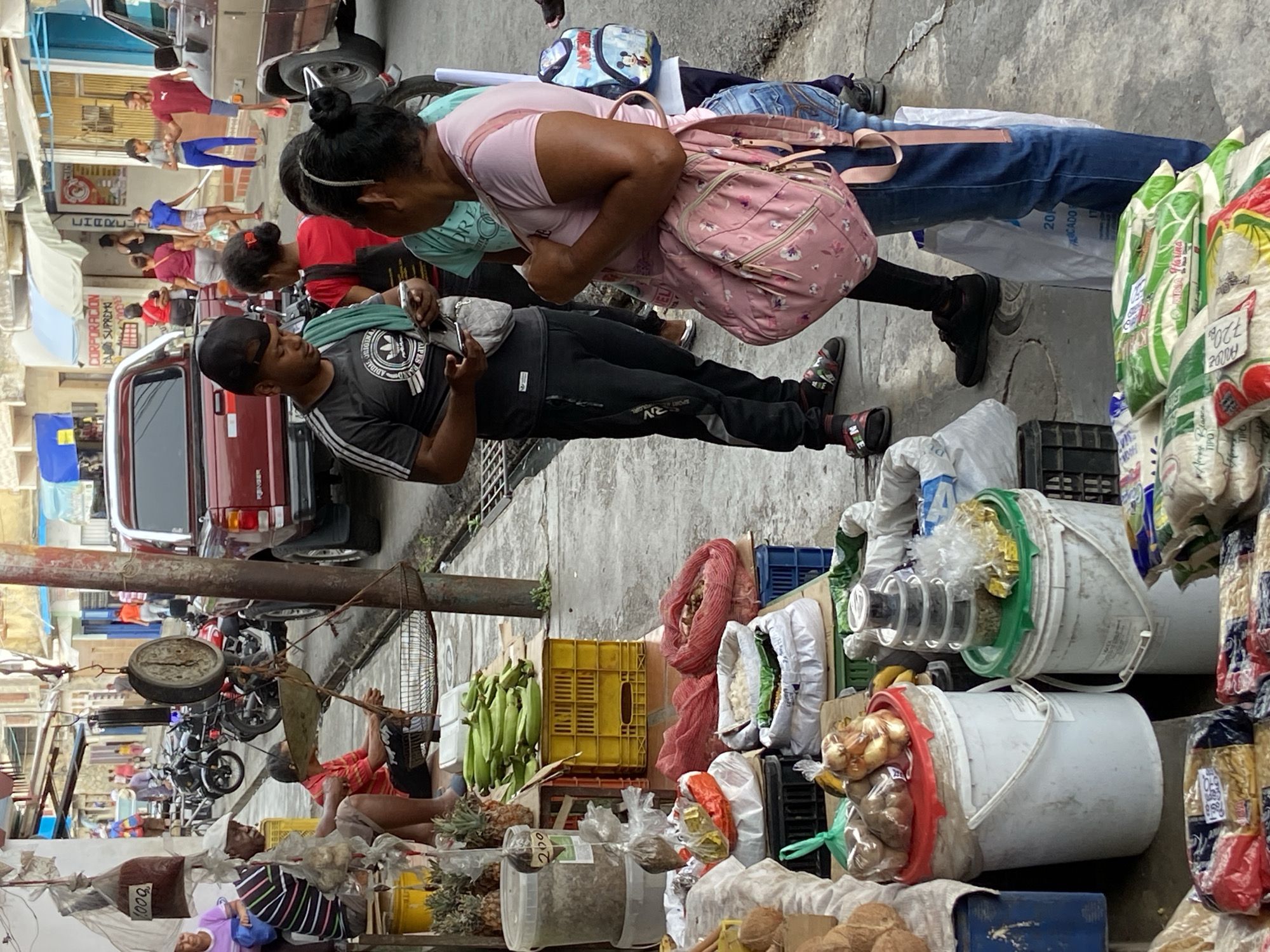 This image captures a vibrant street scene, likely in a market, in what appears to be San José de Barlovento, Venezuela. Several people interact in a crowded outdoor market setting. A man in a black Adidas tracksuit and cap holds an object, potentially money or goods, in his hands, as a woman with a pink floral backpack looks at the object. The road is lined with various goods on display, including fruits like bananas and pineapples, sacks of grain, and onions in buckets. An old-fashioned scale hangs from a metal support structure, behind which a seated man appears to be weighing goods. In the background, several motorcycles are parked. Buildings are visible on the other side of the street, and a red pickup truck parked nearby. The OCR identifies the writing "CORPORACION SUPREMO MYRIGINA" on one of the buildings.