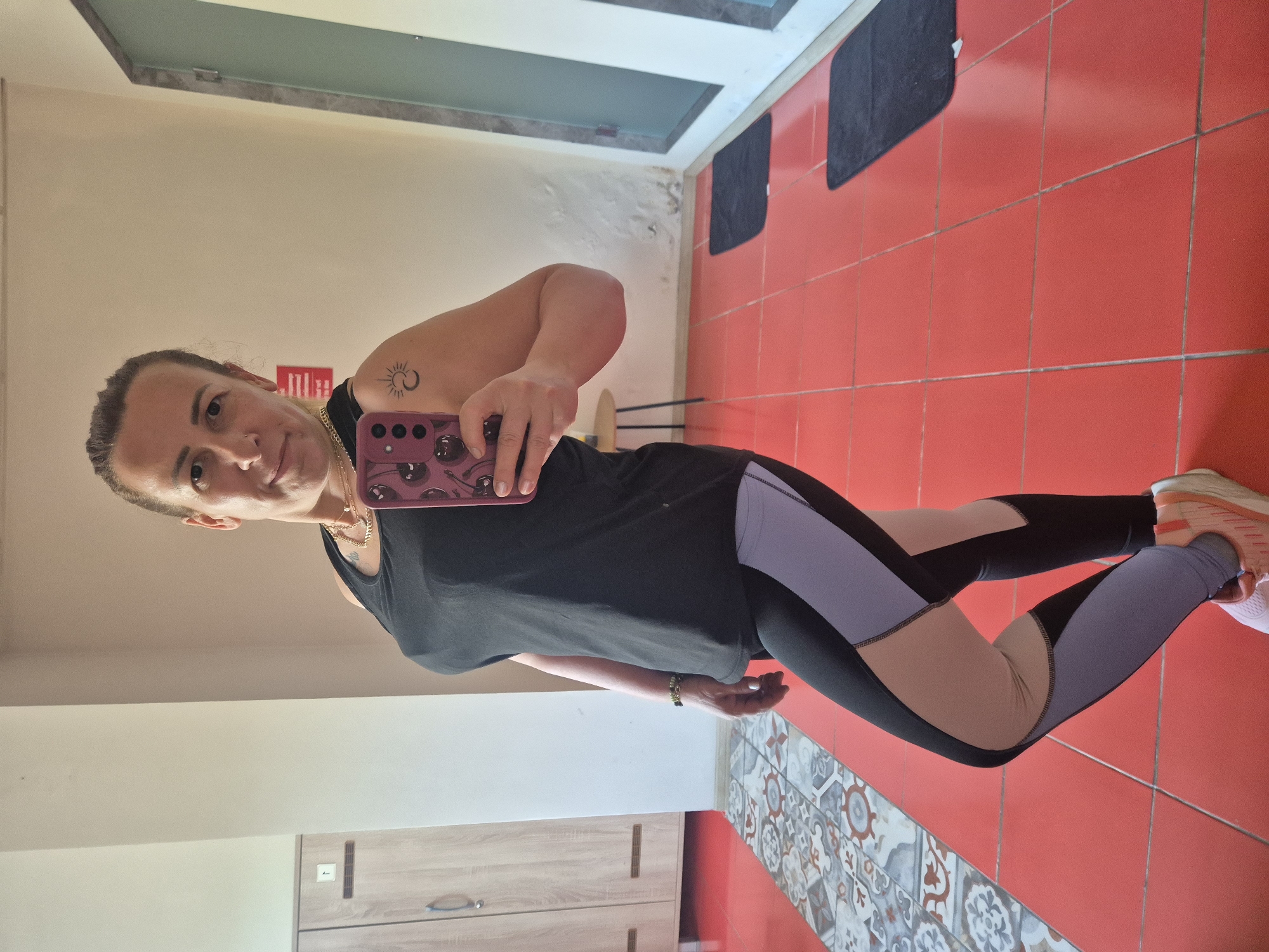 A woman is taking a mirror selfie in what appears to be a locker room or gym. She is wearing a black tank top, multi-colored leggings, and athletic shoes. She has a tattoo of a sun and crescent moon on her left shoulder. In the background, there are lockers, a mirror, a door, and a floor with red tiles and a patterned border. The room is brightly lit.