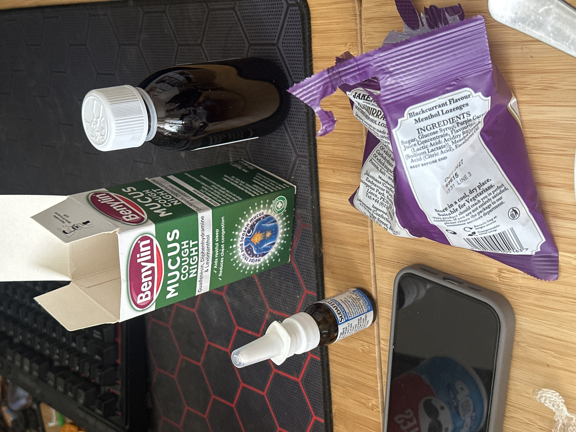 The image shows a collection of over-the-counter cold and flu remedies arranged on a desk. These include an opened box of Benylin Mucus Cough Night medicine, a bottle of liquid cough syrup, Sudafed nasal spray, and an opened package of Jakemans Blackcurrant Menthol Lozenges. A black keyboard and a smartphone with a Pepsi logo are also on the desk. The image was likely taken indoors, and nothing in the image allows identification of a specific location within Wealden or any other place in the United Kingdom.