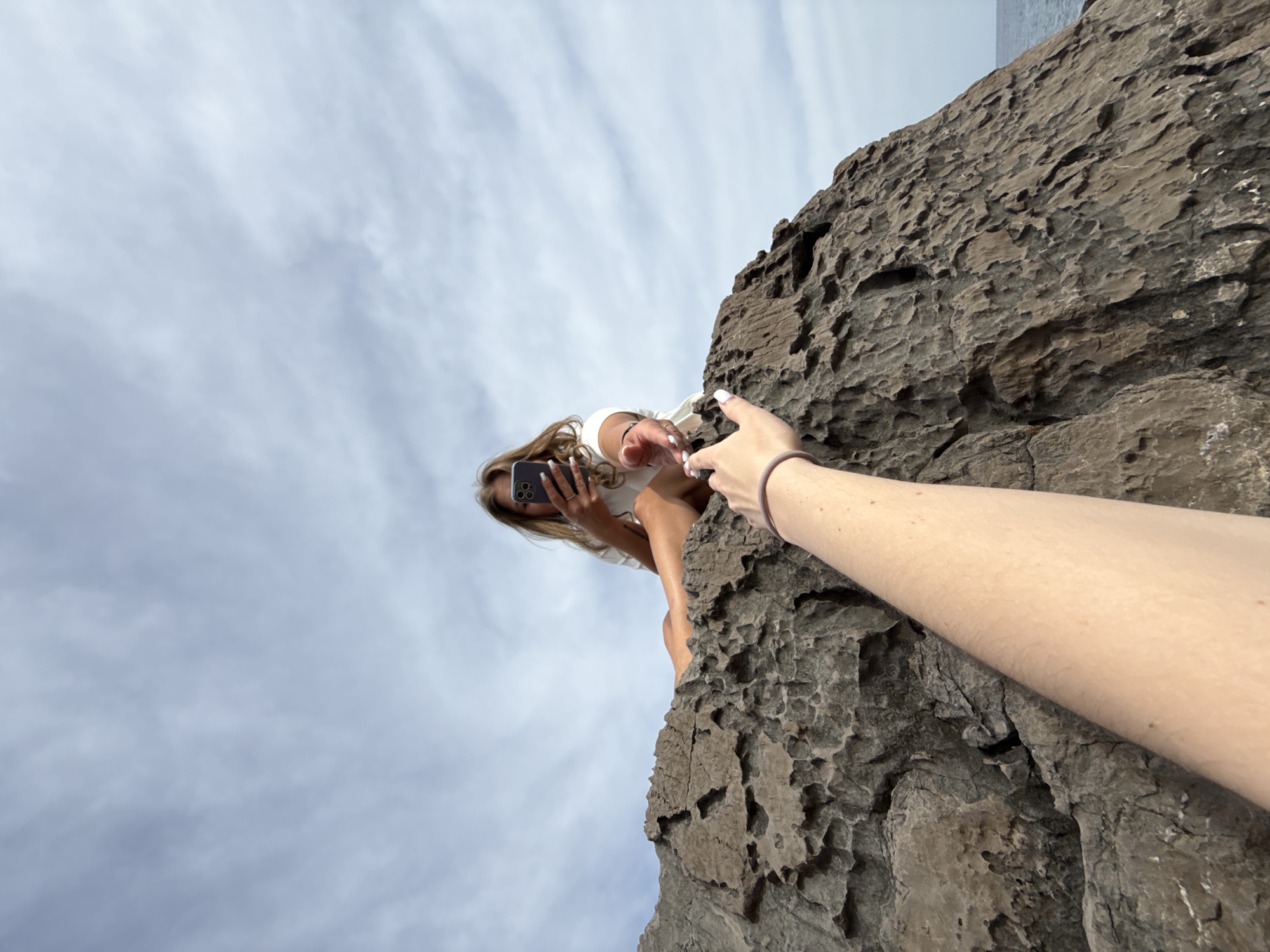A low-angle shot shows a woman sitting on the edge of a dark rock formation under a cloudy sky. She is holding a smartphone and looking at the screen. An outstretched arm with fair skin and a bracelet is extended towards her. The location is Seoca, Montenegro.