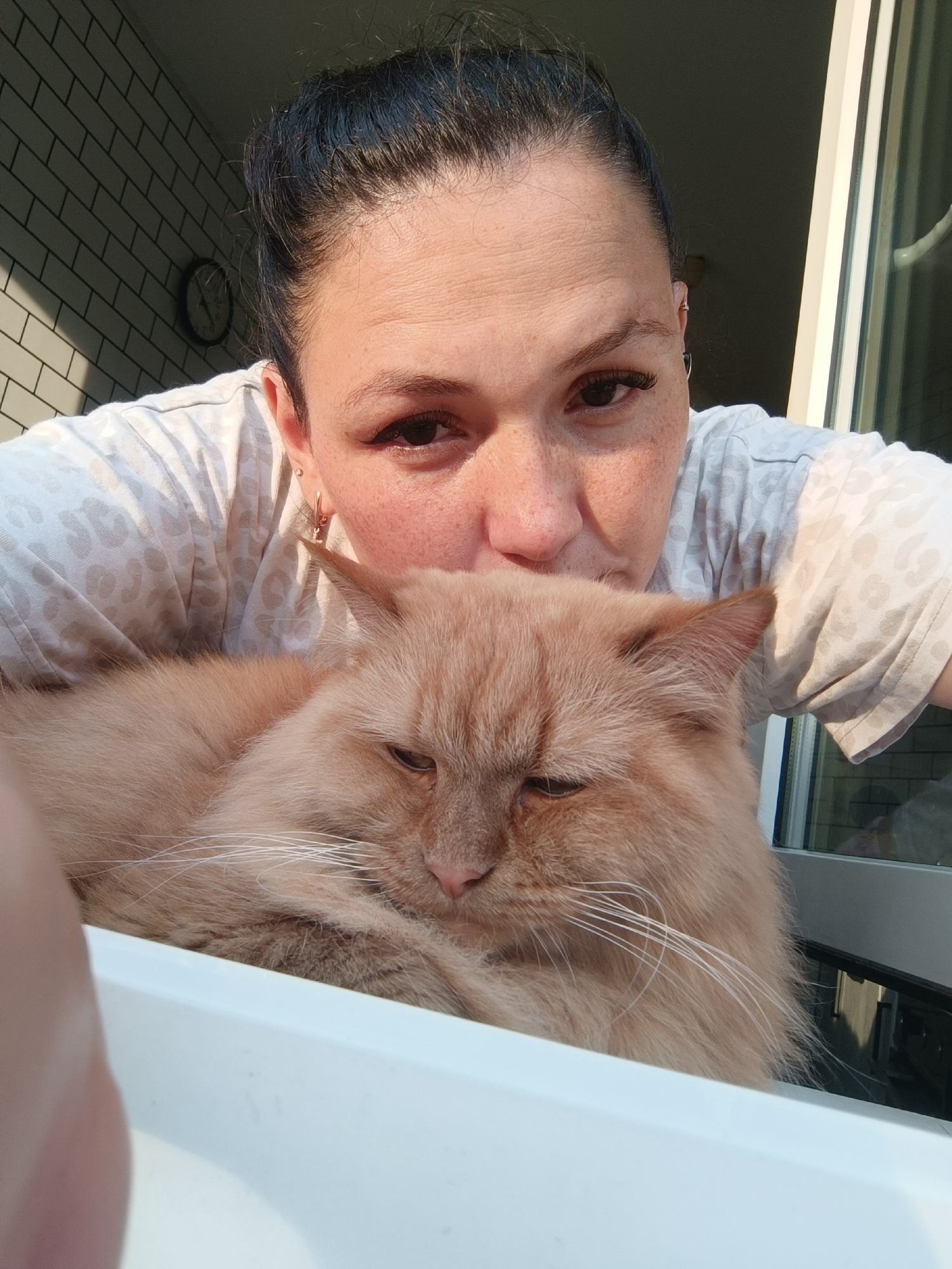 In the city of Chaikovsky, Russia, a selfie is captured of a woman and her ginger cat. The woman is leaning over the cat, with her face close to its head. She has dark hair pulled back and is wearing a light-colored t-shirt with a leopard print pattern. She has freckles and appears to be wearing makeup, including false eyelashes. The cat, a fluffy ginger breed, is resting its head and looking slightly away from the camera, with one eye partially closed. The background shows a white tiled wall with a clock on it, and a window or door with vertical blinds. The scene is illuminated by sunlight, casting shadows. The woman's hand is partially visible in the foreground, suggesting she is holding the camera. The overall impression is an intimate moment between a pet owner and her cat in a domestic setting.