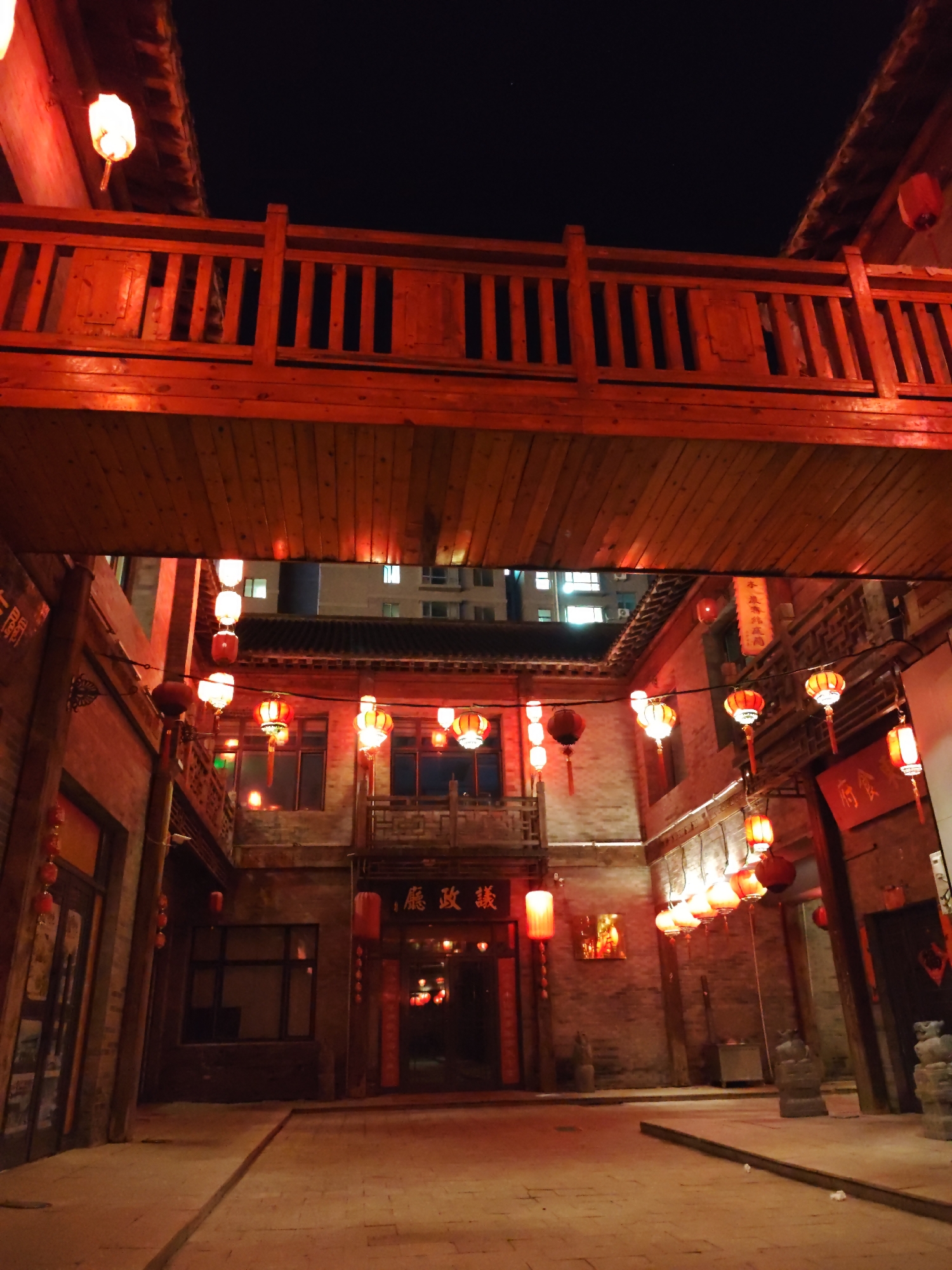 The image depicts a night scene in Lindian, China, characterized by traditional architecture adorned with numerous red lanterns. A wooden bridge, painted red, spans across the top of the frame, with its underside showing wooden planks. Below the bridge, the scene opens into a courtyard surrounded by brick buildings. Red lanterns are strung along wires and hung from the buildings, casting a warm, reddish glow on the surroundings. Some lanterns are spherical, while others are cylindrical. Chinese characters are visible on signs and banners on the buildings. The ground is paved with stone. The overall atmosphere is festive and illuminated by the ambient light of the lanterns against the dark night sky.
