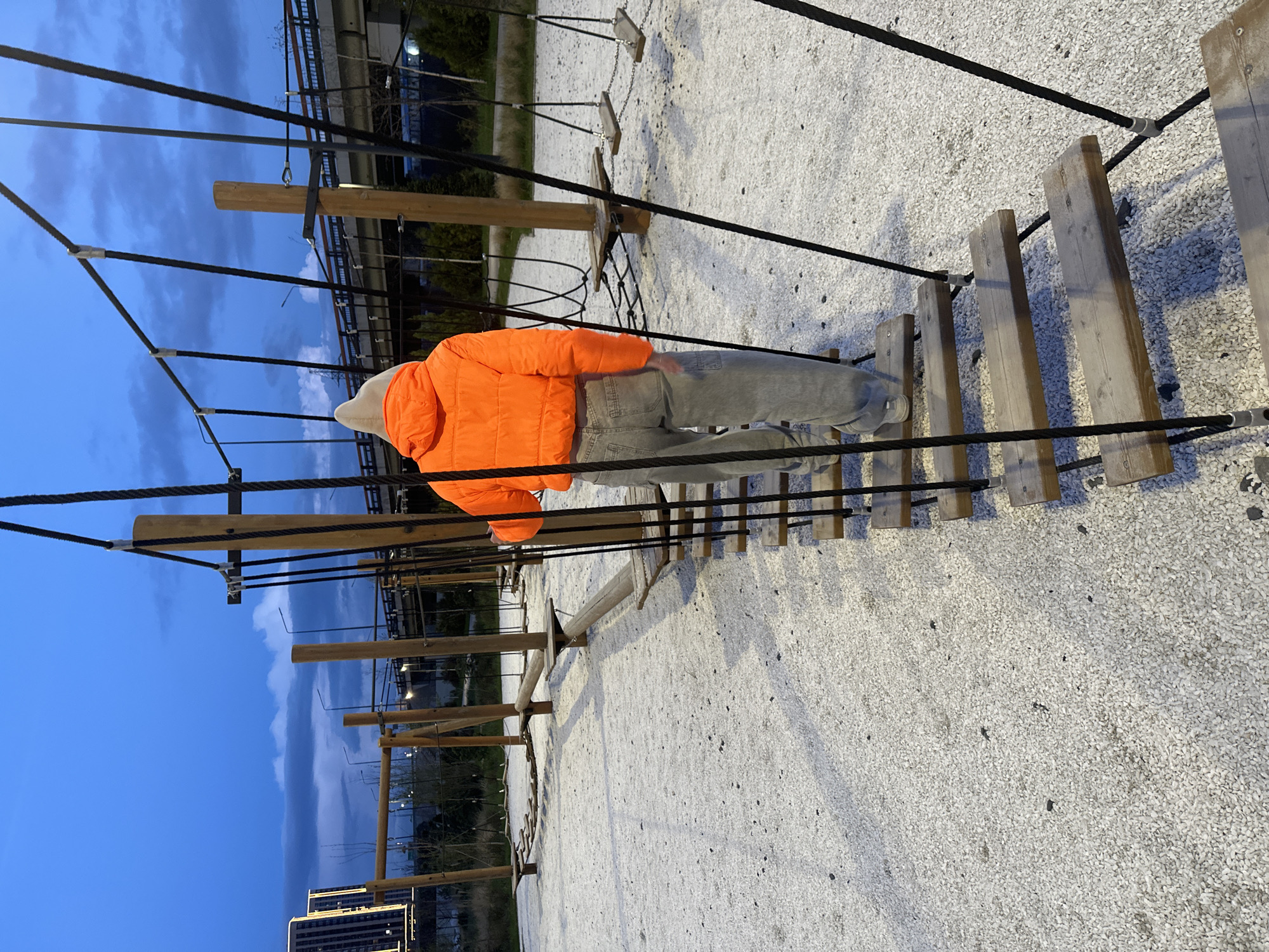 A person wearing an orange jacket and light-colored jeans is navigating a rope bridge obstacle course. The course consists of wooden planks suspended between black ropes and metal supports. The ground is covered in light-colored gravel. In the background, there is a blue sky, a building and a bridge. The image is taken in Rostov-on-Don, Russia.