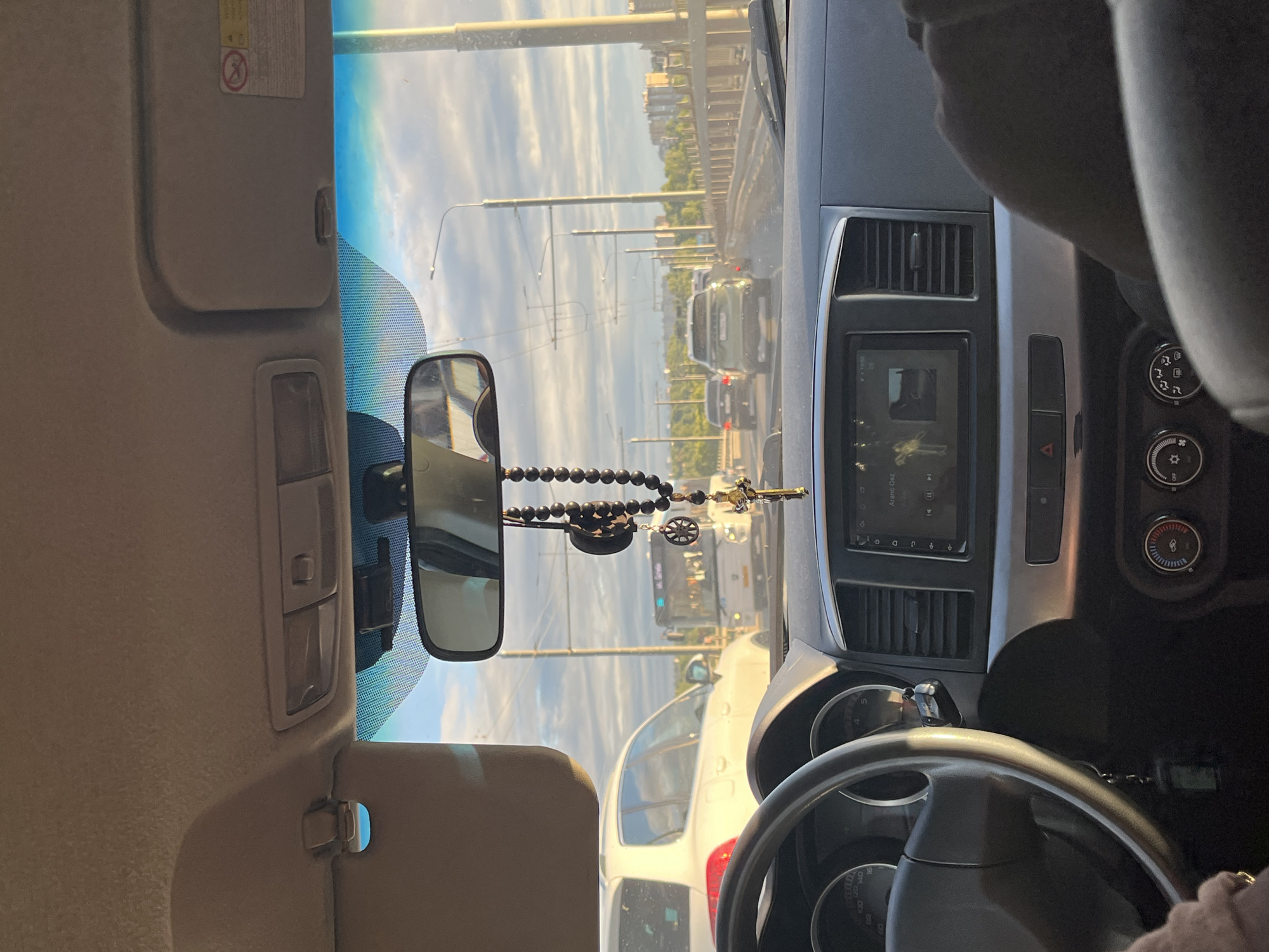 The image shows the interior of a car driving on a bridge, likely over the Volga River in Kostroma, Russia, as suggested by the urban skyline and river visible in the background. The perspective is from the passenger seat, looking forward through the windshield. A black beaded rosary with a crucifix is hanging from the rearview mirror. The dashboard features a multimedia screen displaying what appears to be music playback. Outside, other vehicles, including a white SUV, a bus, and several cars, are visible on the bridge, suggesting moderate traffic. The sky is partly cloudy, with the sun casting a warm light on the scene. The presence of overhead wires indicates the bridge is likely used by trolleybuses or trams.