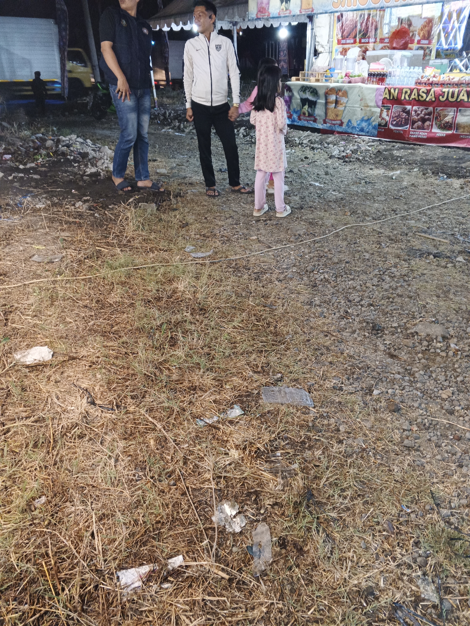 The image captures a nighttime scene at a street food stall in Sukabumi, Indonesia. In the foreground, dry grass and gravel litter the ground, with some plastic debris scattered around. In the midground, three individuals are present. Two men stand by the food stall, one wearing a dark t-shirt and jeans, and the other in a white jacket and black pants. A young girl, dressed in a pink outfit, stands facing the stall, her back to the camera. The food stall itself is illuminated and displays various food items and beverages. Behind the stall, part of a white truck and a yellow vehicle are visible. The overall environment suggests a bustling, open-air market setting.