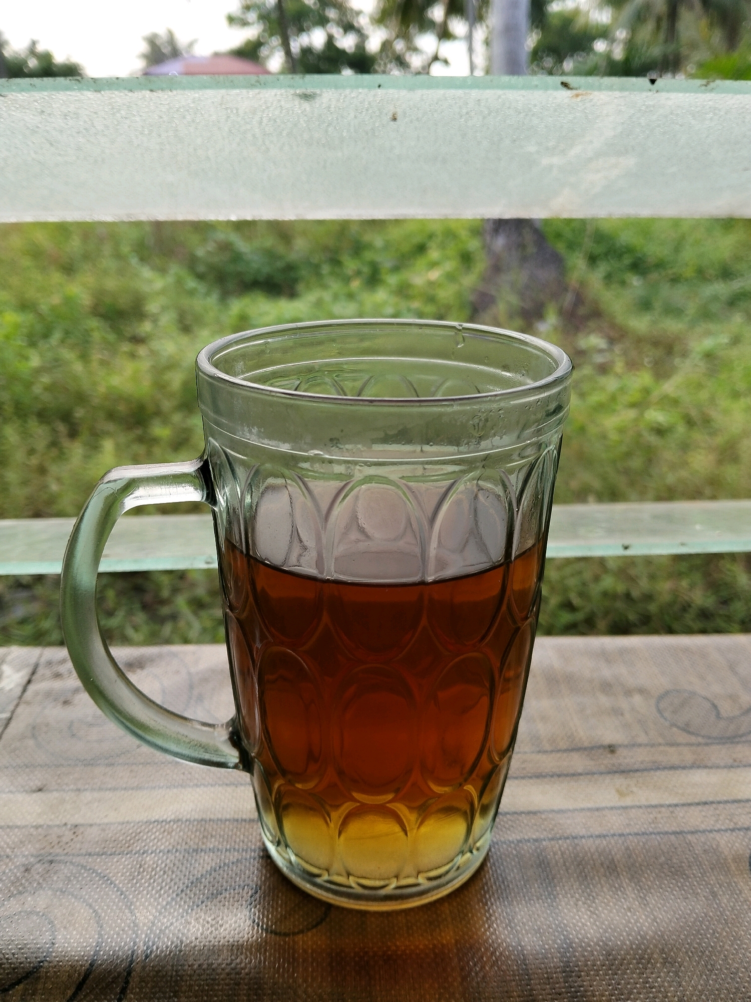 A close-up, eye-level shot shows a glass mug filled with dark amber liquid and a lighter yellow substance at the bottom, possibly tea and honey, sitting on a patterned surface. The mug has a clear glass handle and decorative textured patterns on its body. In the background, out of focus, is lush green vegetation and trees, suggesting an outdoor setting. A frosted translucent bar runs horizontally across the upper portion of the frame, with faint greenery visible beyond it. The scene is set at a table outdoors, with the humid, tropical ambiance of Teluk Pambang, Indonesia, evident in the soft, diffused light and abundant foliage. There are no people or animals present in the image.