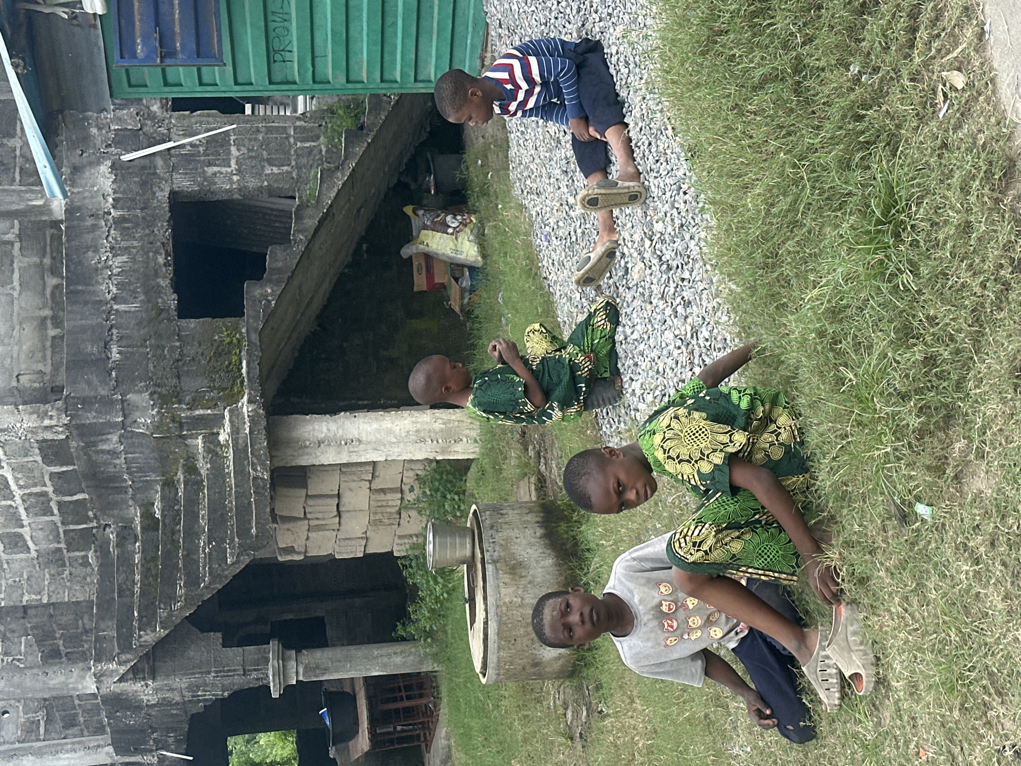 Four young children are seated on the ground near an unfinished concrete building with a prominent outdoor staircase in Badagry, Nigeria. The building appears to be constructed from rough-hewn stone blocks and concrete. To the right of the building is a bright green structure with a blue roof, possibly a shipping container or a small kiosk.
In the foreground, two boys are sitting on a patch of grass. One boy, wearing a gray t-shirt with cartoon cat faces and dark pants, is looking towards the left. Beside him, another boy in a green and yellow patterned traditional outfit is looking at the viewer.
Further back and to the right, two more boys are seated on a gravel surface. One boy is wearing a green and yellow patterned outfit, similar to the one described above, and appears to be holding something in his hands. The other boy is wearing a blue and white striped long-sleeved shirt and dark pants, with his legs extended.
Behind the children, a circular concrete structure resembling a well or water cistern is visible. A metal bucket sits on top of it. The environment is somewhat overgrown with grass and weeds, and debris is scattered on the ground. The overall scene suggests a casual gathering of children in an outdoor area adjacent to a partially constructed building.