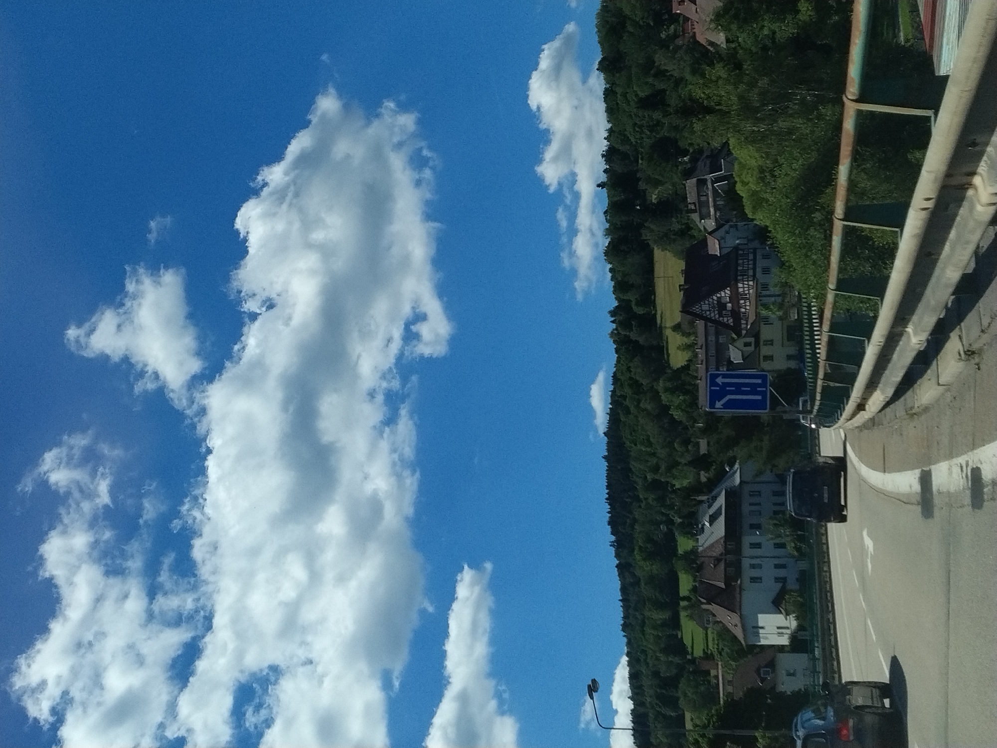 The image captures a view from a vehicle on a road approaching a town nestled amongst forested hills. The sky is a bright blue with scattered white clouds. The road curves to the right, with a metal guardrail on the right side. Several cars are visible on the road, including a black van in the foreground and a dark-colored car on the left.
In the background, a cluster of buildings forms a small town. One prominent building features a traditional half-timbered architectural style with a red roof. Adjacent to it is a larger, light-colored building with multiple windows. The town is surrounded by lush green trees that extend up the hillsides.
A blue traffic sign with white arrows indicating lane direction is visible on the right side of the road. A street lamp is also present on the left side of the road, near the crest of the hill. The overall scene depicts a scenic drive through a rural European landscape, leading into a village or small town. There is no clear indication that this location is specifically Vimperk, Czechia, as there are no identifiable landmarks or text to confirm the location.