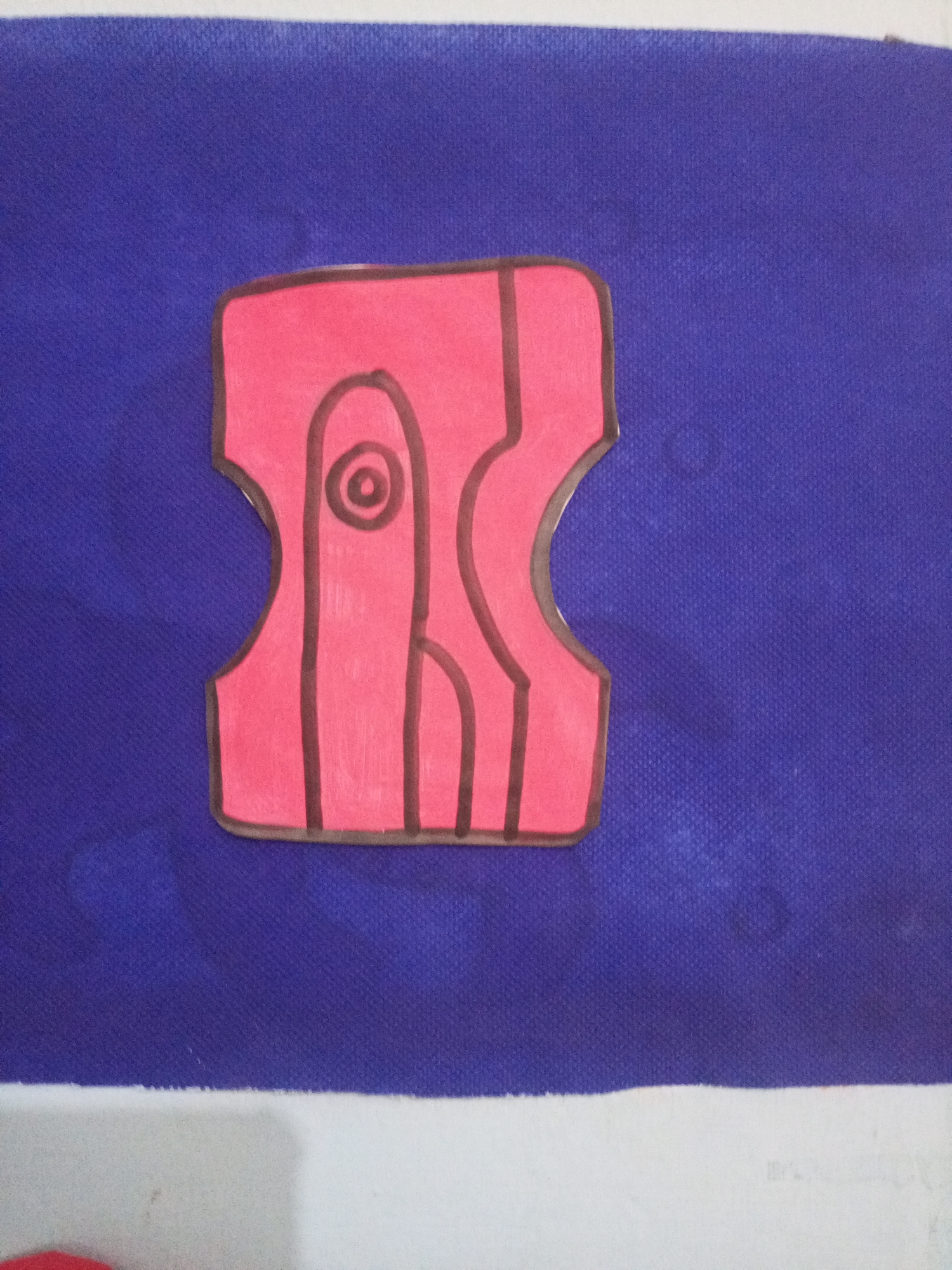 A drawing of a red pencil sharpener with black outline is placed on a blue textured surface. The pencil sharpener is stylized and abstract, with internal lines that suggest the opening for the pencil and the blade. The drawing appears to be a children's artwork. The background is a solid blue fabric, and a white surface is visible at the bottom of the image. There are no identifiable landmarks or contextual clues within the image that would allow for determining its location in Santa Ana, Venezuela.