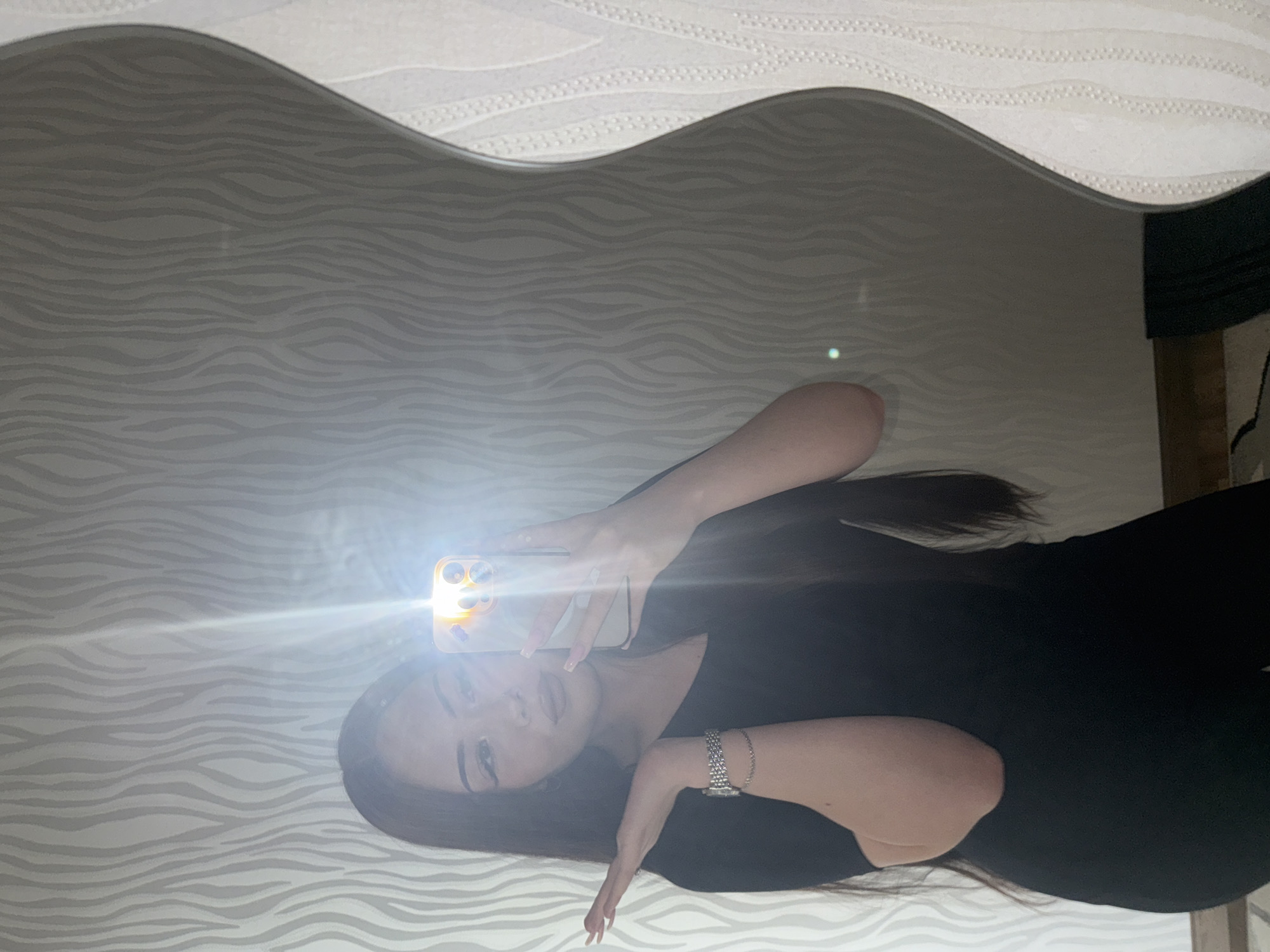 A person is visible in a selfie taken in a mirror. The mirror's frame has an undulating, wave-like design. The background reflects a wall with a pattern resembling horizontal zebra stripes. The subject is holding a smartphone, using the flash. The person is wearing a dark garment and a bracelet on their left wrist. The location cannot be determined to be Naberezhnye Chelny, Russia, or any specific place.