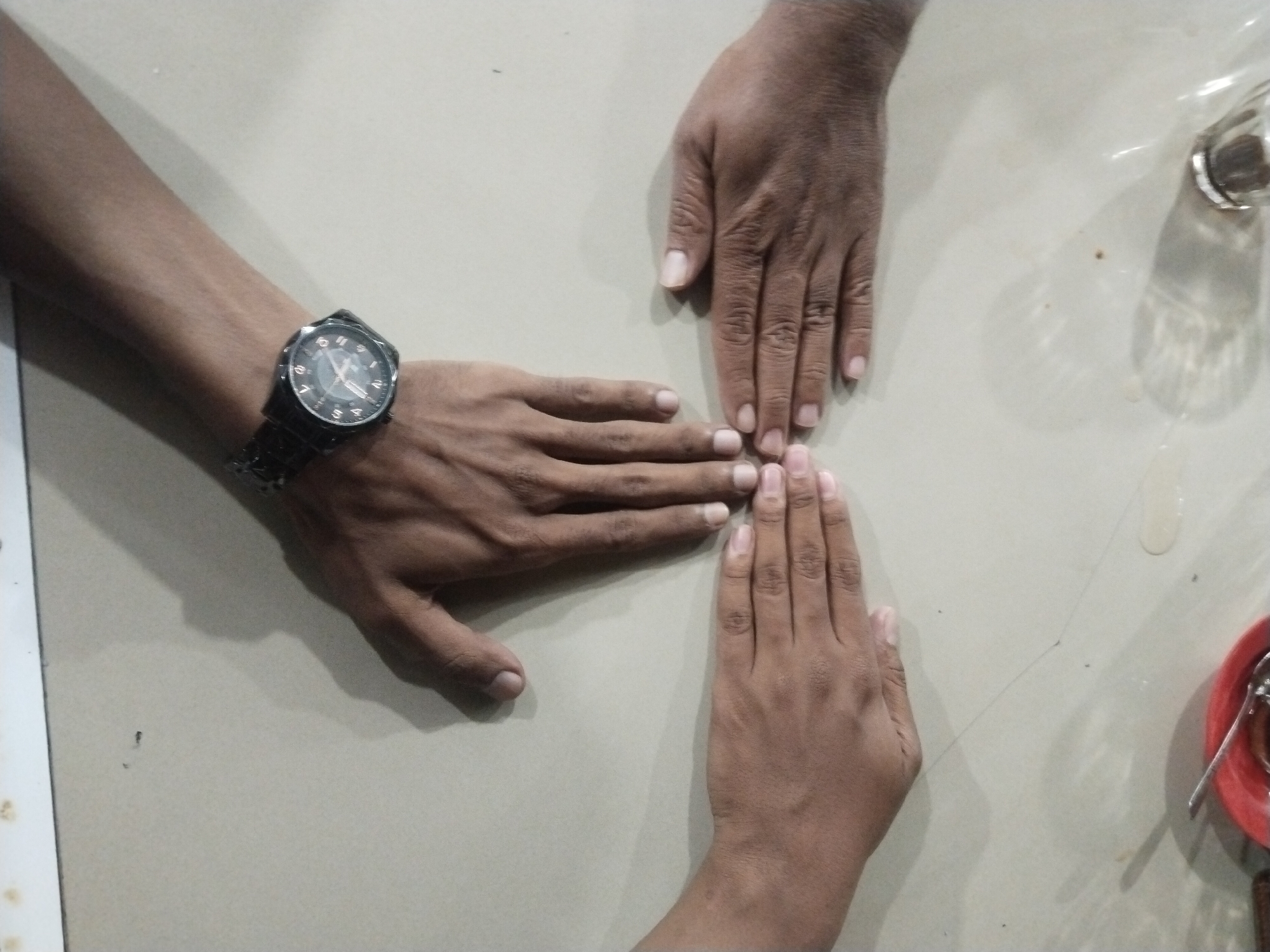 The image shows three hands touching at the fingertips on a light-colored surface. One of the hands has a watch on the wrist. A glass and a red plate with silverware are visible in the upper right corner. The location and city cannot be determined from the image. The activity seems to be casual.