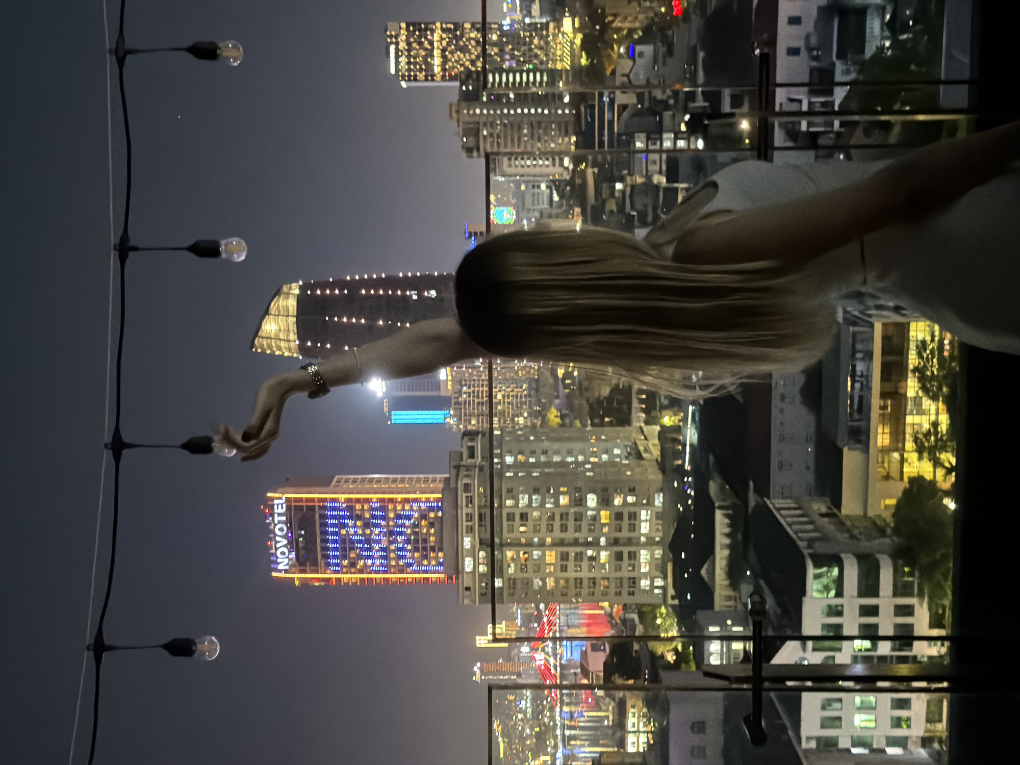 A young woman stands on a balcony at night, her right arm raised, holding what appears to be a small object. She faces away from the camera, looking out at a city skyline. The city, identified as Đà Nẵng, Vietnam, is illuminated by numerous lights from buildings and bridges. A prominent skyscraper in the background displays the "NOVOTEL" logo. String lights with exposed bulbs hang across the top of the frame. The woman has long, blonde hair and wears a white sleeveless top and a patterned skirt. The balcony railing is made of glass.