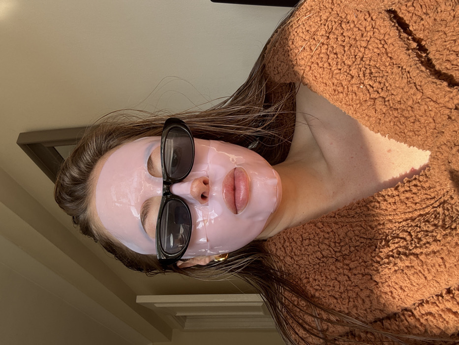 A female subject is depicted indoors, likely within a room or hallway, in the city of Unknown, Unknown. She is centered in a medium close-up, looking directly at the camera. Her face is covered by a translucent pink hydrogel sheet mask, with cutouts for the eyes, nostrils, and mouth. Black, oval-shaped sunglasses are worn over the mask, obscuring her eyes. Her lips are visible through the mask's mouth cutout. Long, damp-appearing dark brown hair frames her face and shoulders. A single, large, gold-colored hoop earring is present on her left ear. She is clad in a thick, textured, rust-brown garment, possibly a robe, open at the neckline. The background features a light-colored wall, a section of a white doorframe or molding to the upper left, and a dark-framed object, possibly a mirror or picture, to the upper right. The scene is illuminated by strong, warm-toned natural light from the left.