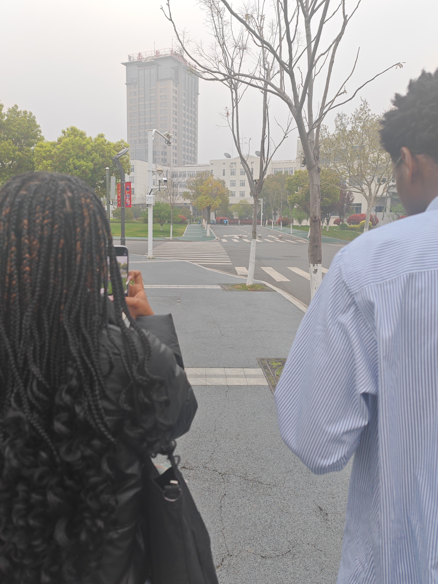 The image shows a street scene in the Jiangning District, China. Two individuals are visible, a woman with long braids is taking a photo with her cell phone, and a man in a blue striped shirt is standing beside her. The street has crosswalks, trees, and light posts. In the background, there's a tall building and other structures, all partially obscured by a hazy atmosphere.