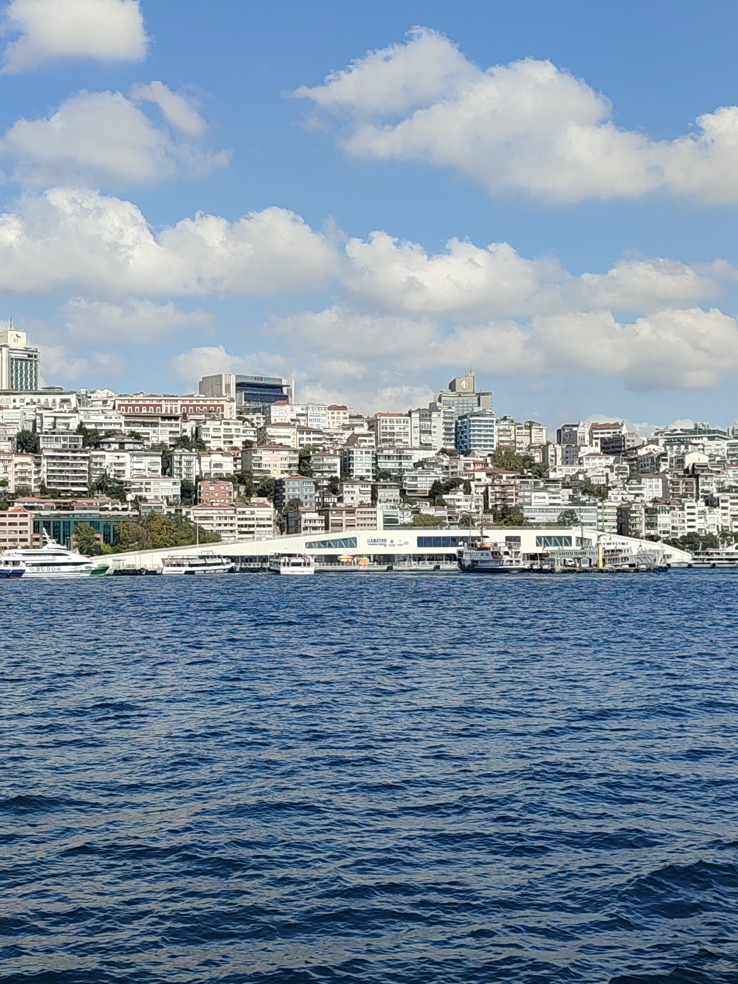 The image captures a panoramic view of the Beyoğlu district in Istanbul, Turkey, as seen from the Bosphorus. The foreground is dominated by the deep blue, rippling waters of the strait, reflecting the bright sky above. Several vessels are visible on the water, including ferries and a white yacht with "BUDO" written on its side, suggesting passenger or transportation services.
In the mid-ground, a modern waterfront structure, likely a ferry terminal, runs along the coastline. The building features a long, white facade with large glass windows and a covered walkway. Behind this structure, the densely populated Beyoğlu district rises up a steep incline. The cityscape is characterized by a dense collection of buildings, predominantly white and light-colored, with varying architectural styles. Many structures appear to be apartment buildings or offices, with a mix of modern and older designs. Some taller buildings stand out amongst the lower-rise structures, including a prominent skyscraper with a circular clock face near its summit. The hillsides are dotted with trees and greenery, interspersed between the buildings.
The sky above is a vibrant blue, punctuated by fluffy, white cumulus clouds. The sunlight suggests a clear and pleasant day, casting shadows and illuminating the scene. The overall impression is of a bustling urban environment situated on a picturesque waterfront.