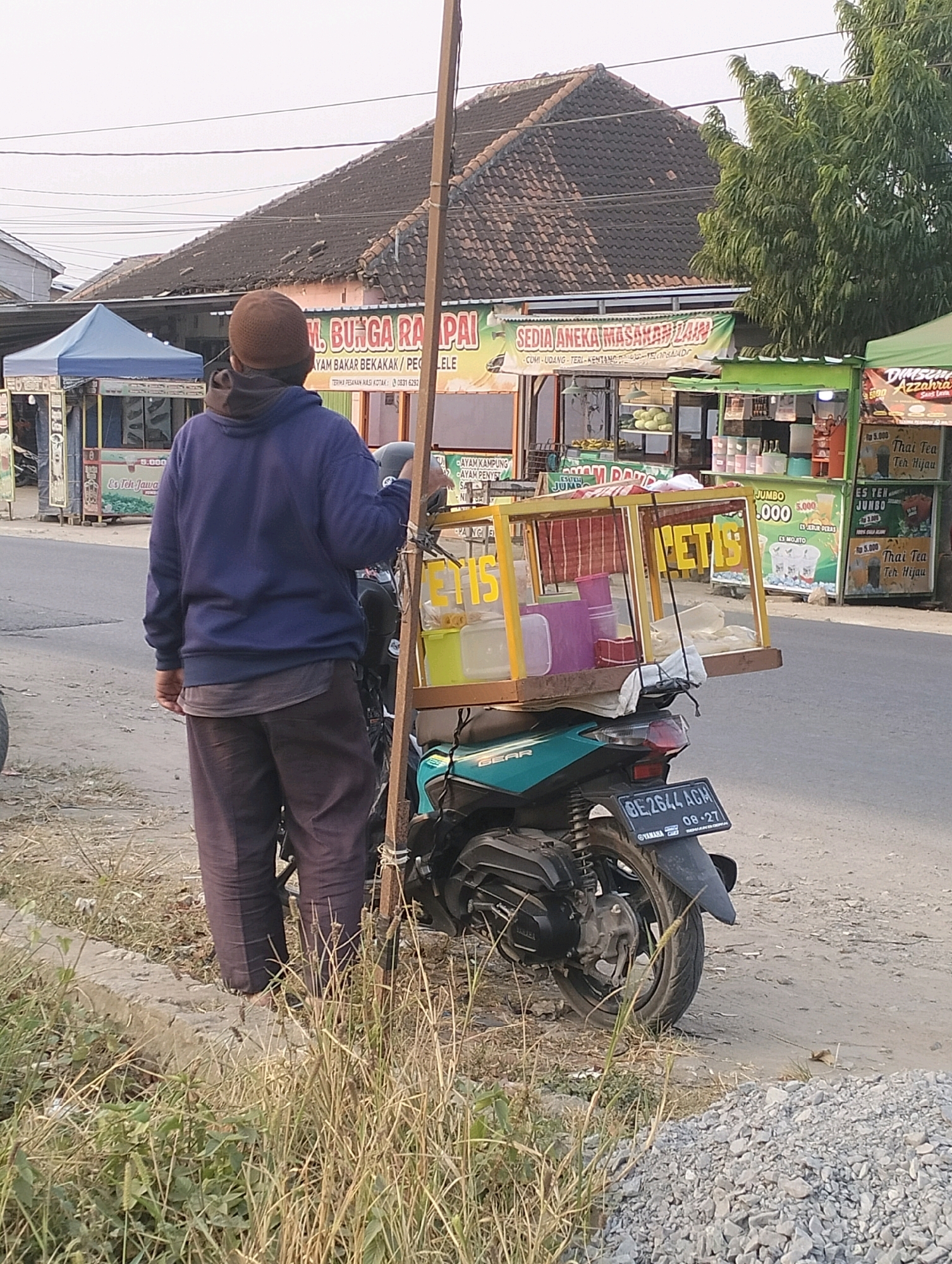 A person wearing a blue hoodie and dark pants stands next to a teal motorcycle with a mobile food cart attached. The food cart is made of glass and wood and contains various containers, suggesting it is used for selling food or drinks. The motorcycle has a license plate that reads "BE 2644 AGM". In the background, there are several roadside stalls with awnings and signs advertising food items like "AYAM BAKAR BEKAKAK" and "SEDIA ANEKA MASAKAN LAIN". The scene appears to be a street or roadside in a commercial area, likely in Perumnas Way Kandis, Indonesia, given the context. The sky is overcast, and the ground is a mix of dirt and gravel. There is a wooden pole attached to the motorcycle, extending upwards, possibly for display or support of the food cart. The overall impression is of a typical street vendor scene in a bustling urban environment.