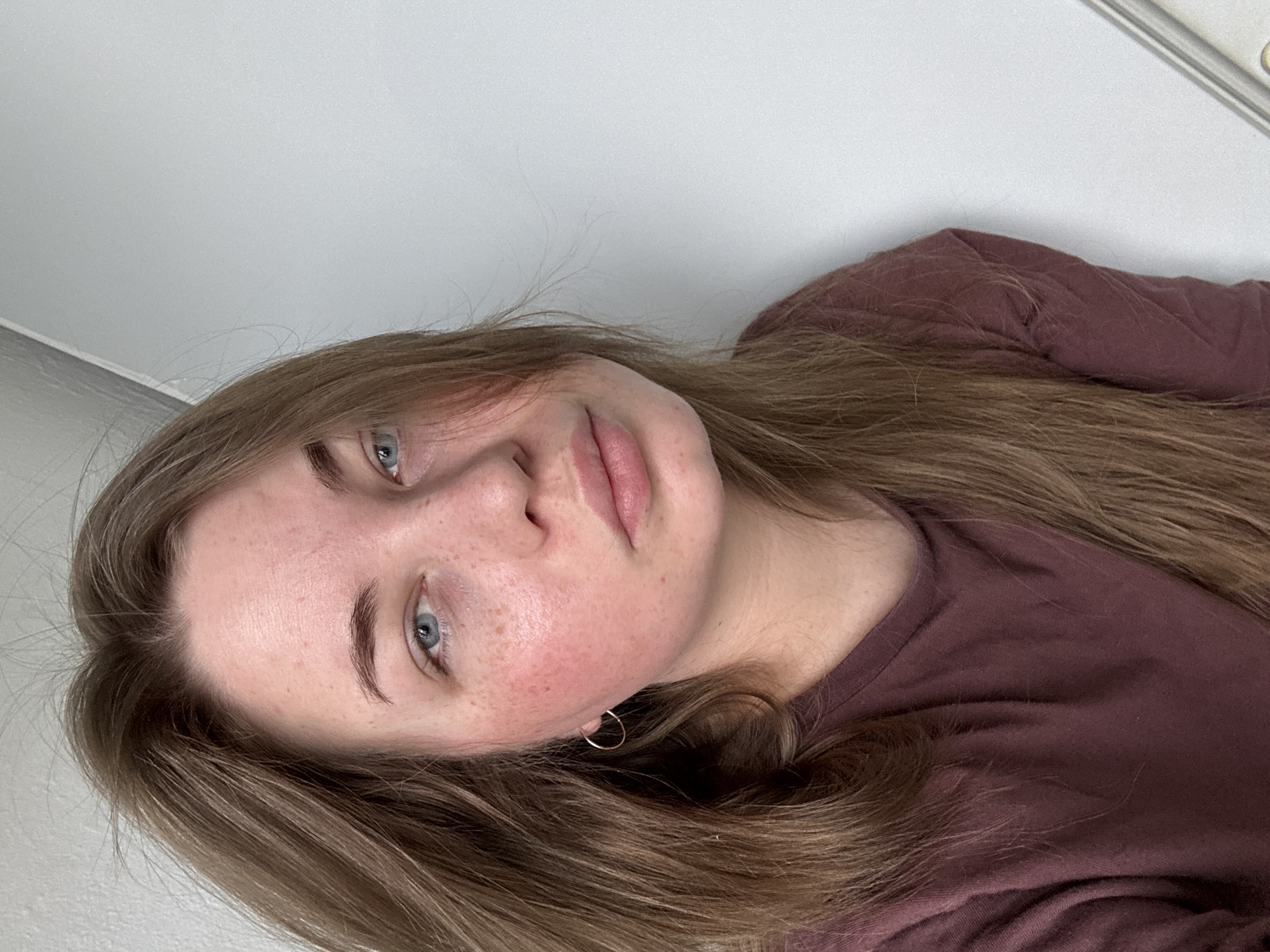 A woman with light brown hair, blue eyes, and fair skin with freckles is positioned at an angle, facing the camera. She is wearing a maroon t-shirt and a gold hoop earring. The background consists of a plain white wall. There is no indication of location or specific information relating to the city of Chaikovsky, Russia.