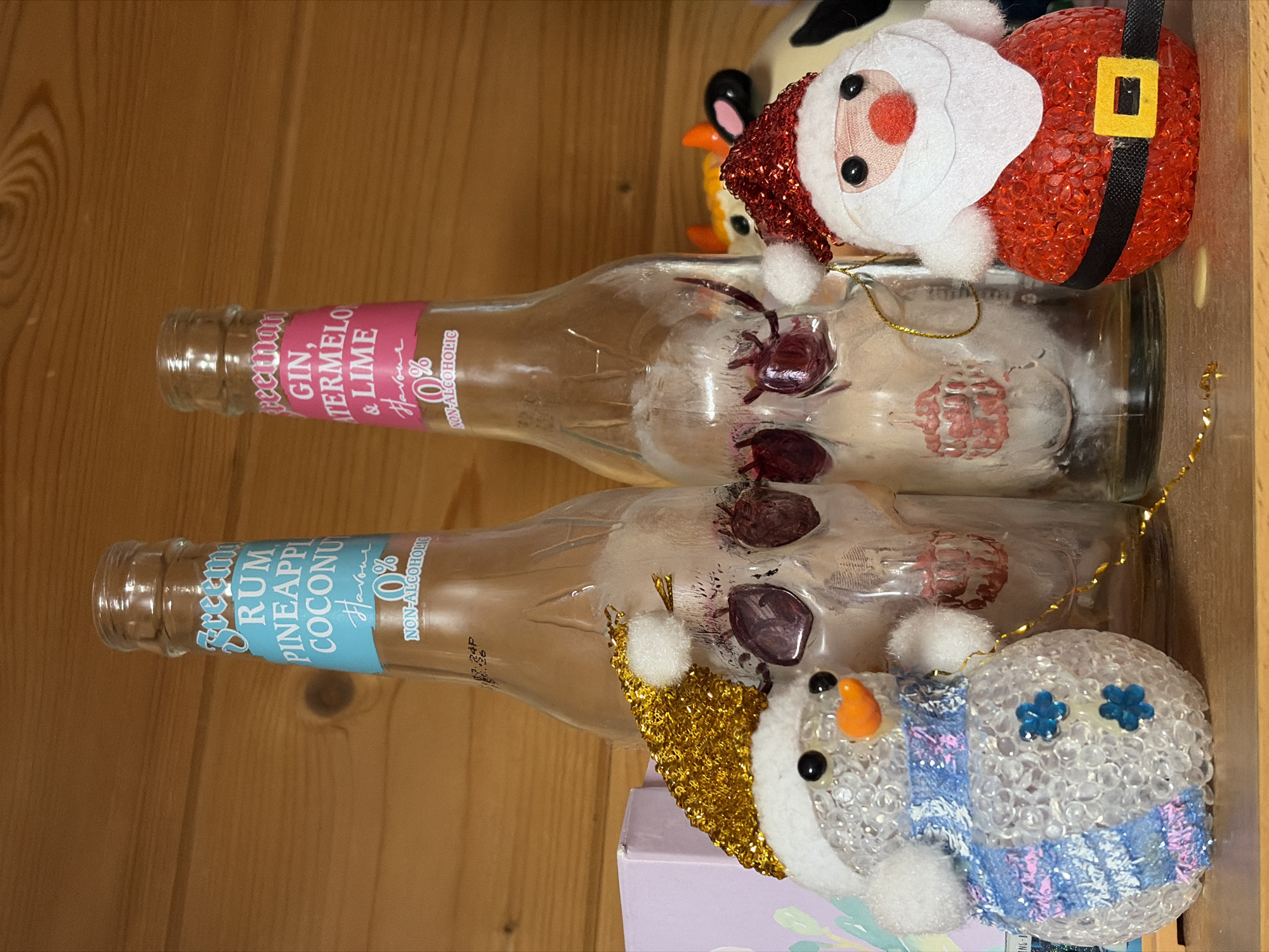 The image presents a close-up, horizontally oriented view of two non-alcoholic beverage bottles and two Christmas ornaments, arranged on a warm, light brown wooden surface. The setting appears to be indoors, potentially in Прикамский, Russia, although no specific location cues are visible.
The top bottle, clear glass, features a pink label. Visible text on the label reads: "Freeman GIN, WATERMELO & LIME Flavour 0% NON-ALCOHOLIC". Inside the bottle, there are dried, dark red or purple residues. Resting on this bottle is a Santa Claus ornament, characterized by a red glittery body, a black belt with a gold buckle, a white felt beard and hat trim, a red nose, and black bead eyes.
The bottom bottle, also clear glass, has a light blue label. The text on this label states: "Freeman RUM PINEAPPL COCONU Flavour 0% NON-ALCOHOLIC". Similar to the top bottle, dark residues are visible inside. Beside this bottle, positioned on a partially visible light purple box, is a snowman ornament. It is constructed from clear beaded material and features a white hat with gold glitter, an orange carrot nose, black bead eyes, and a blue and pink striped scarf. Two small blue flower details adorn its head.
The overall scene suggests a festive or decorative display, likely related to the Christmas holiday due to the ornaments. There is no activity taking place, and the items are static. The bottles appear to be empty or contain only dried remnants of their original contents.