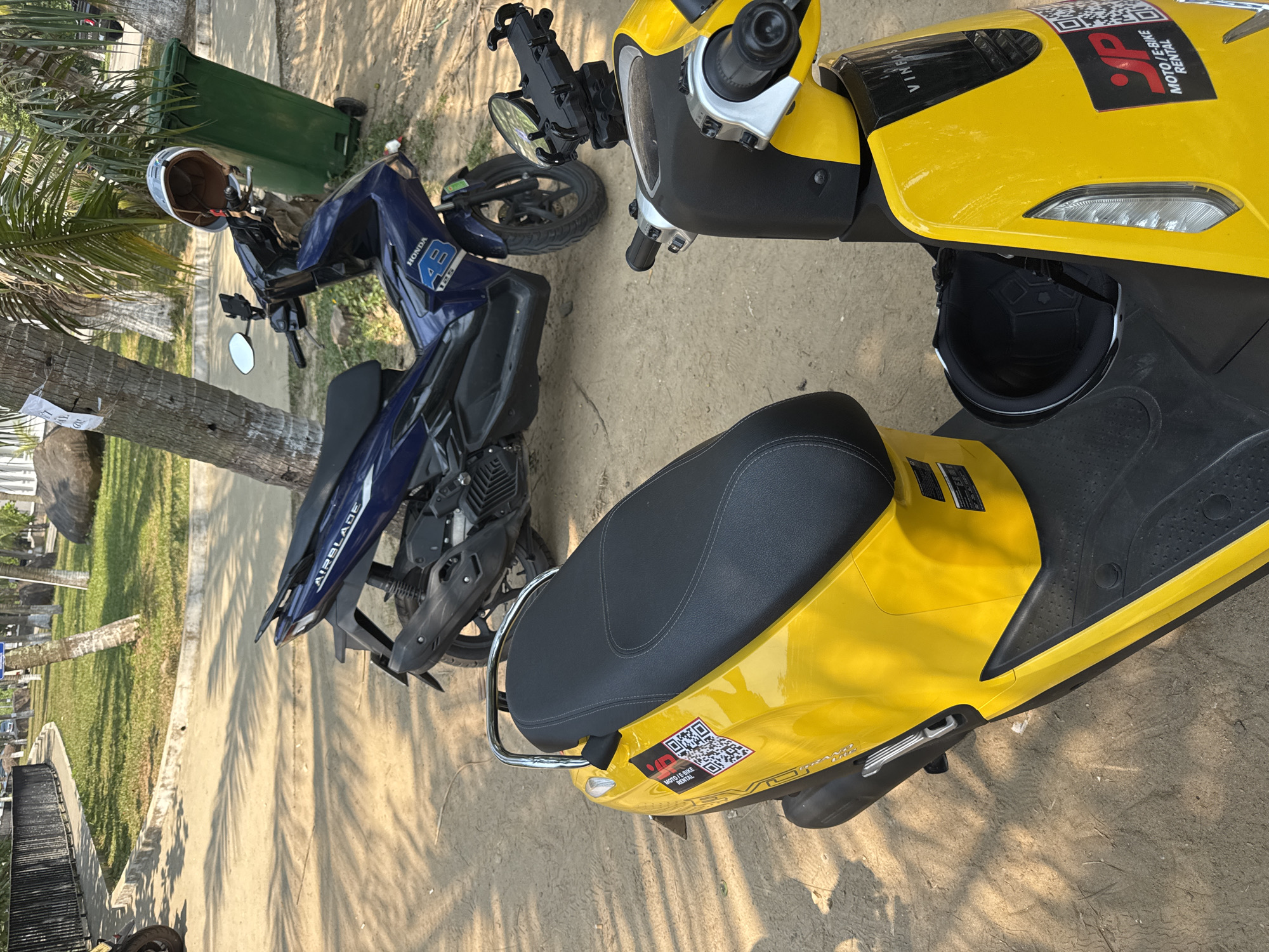 The image shows two parked motorbikes on a dirt and concrete surface. The closest motorbike is yellow and has a black seat. It has stickers indicating "VinFast" and "MOTO/E-BIKE RENTAL." The motorbike beside it is blue and appears to be a Honda AirBlade 125. A helmet is resting on its handlebars, next to a green trash bin. A palm tree is visible to the left, casting shadows on the ground. The setting appears to be in Đà Nẵng, Vietnam, based on the motorcycle brands and models common in the area. A glimpse of grass and other trees can be seen in the background.