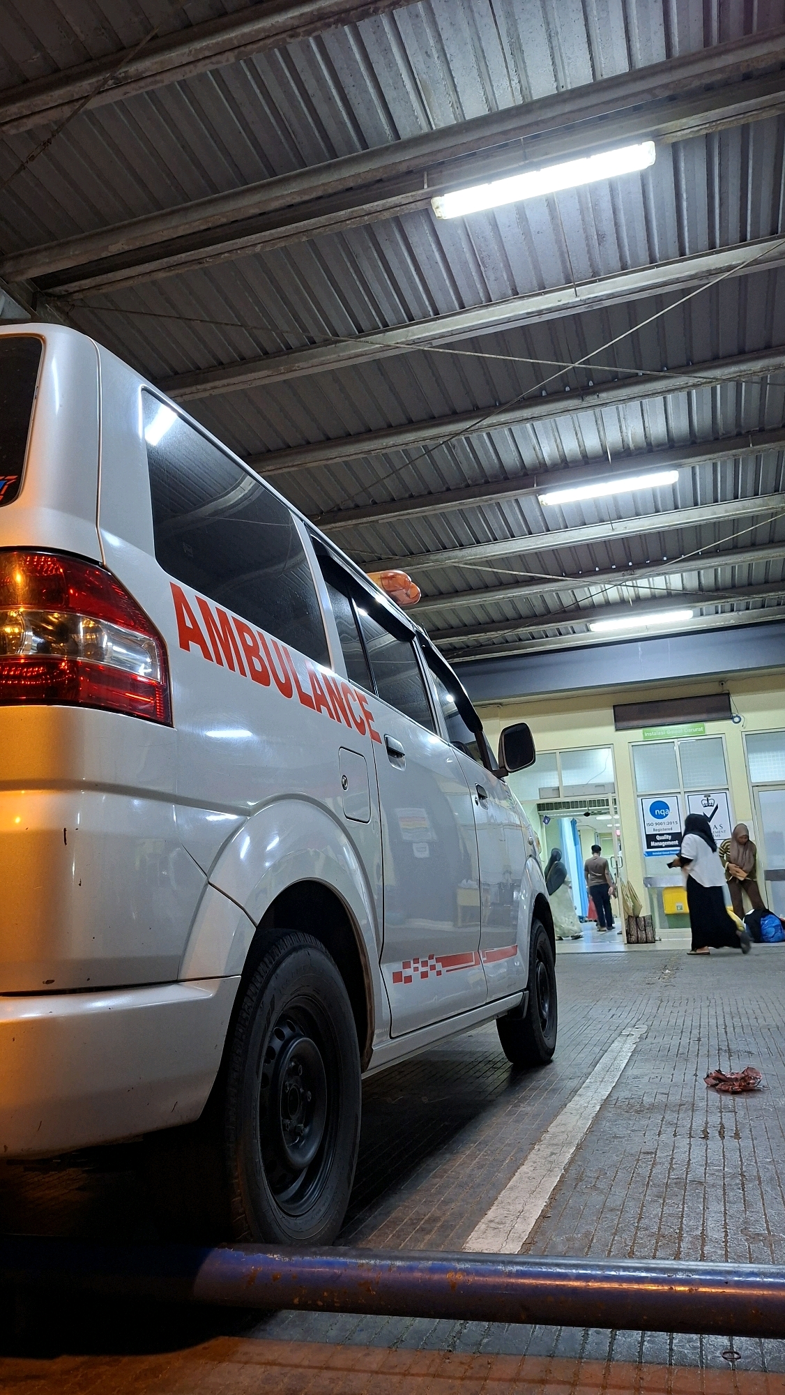 A white ambulance with red "AMBULANCE" lettering is parked on a textured gray tiled surface, presumably at a medical facility. The ambulance is positioned at an angle, with its rear on the left side of the frame and its side visible. A red and white checkered stripe runs along the lower side of the ambulance. The scene appears to be an indoor or covered area, indicated by a corrugated metal roof supported by metal beams and illuminated by fluorescent lights. In the background, there is a building entrance with a green facade and glass doors. Several individuals are visible in the background, including two women in headscarves and one person in a dark shirt and pants. One woman appears to be holding a bag, and another is walking away. The ground also shows some debris or discarded items. The overall atmosphere suggests a functional, perhaps busy, medical environment in Banda Aceh, Indonesia.
