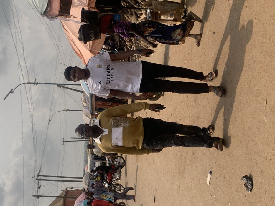 The scene depicts a bustling open-air market in Otuocha, Nigeria, under a bright, overcast sky. Two adult males occupy the foreground, walking towards the viewer's right. The individual on the left wears a gold-yellow long-sleeved polo shirt with a white horizontal stripe featuring "VOLUF" text, dark trousers, and brown sandals; he holds a dark smartphone in his right hand. The individual on the right wears a white short-sleeved t-shirt with an Adidas logo, Real Madrid crest, and 'Emirates FLY BETTER' text, black trousers, and gold-brown sandals; he holds a dark smartphone in his left hand.
The ground is packed earth, with a clear plastic bottle and a crumpled dark plastic bag visible as litter in the foreground. In the mid-ground on the right, an adult female with a red head covering walks past market stalls, balancing two dark buckets marked 'EMEFIA 2' on her head. These stalls are covered by tarpaulin canopies (one orange, one blue) and display numerous piles of root vegetables, likely yams, alongside sacks and a silver tray on a wooden stool.
In the left mid-ground, a red motorcycle is parked, and various individuals are seen walking. The background features several auto-rickshaws and a white van. Numerous utility poles, overhead power lines, and two solar-powered streetlights dominate the upper section of the image. Distant buildings and a hazy horizon are visible. Shadows are cast by the foreground figures, indicating a strong light source.