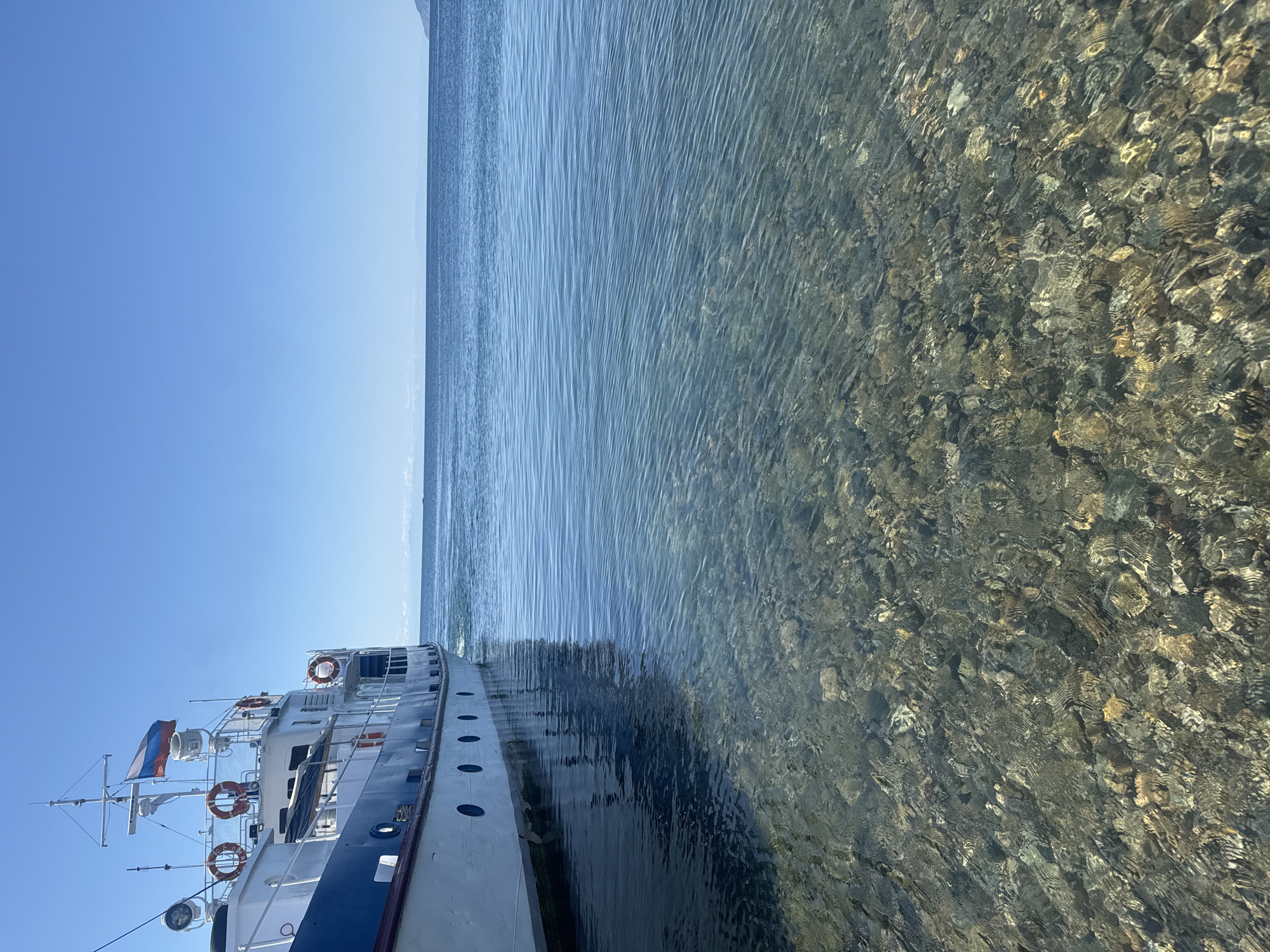 A white and blue passenger boat is docked in shallow, clear water on a sunny day. The boat is positioned on the left side of the frame, with its hull partially submerged. The water is so transparent that the rocky, pebble-strewn bottom is visible. Ripples on the water's surface create a shimmering effect. In the background, a calm, deep blue body of water stretches to the horizon, meeting a clear blue sky. A distant, hazy shoreline with mountains can be seen faintly on the horizon. The Russian flag is visible flying from the boat's mast. The scene is calm and serene.