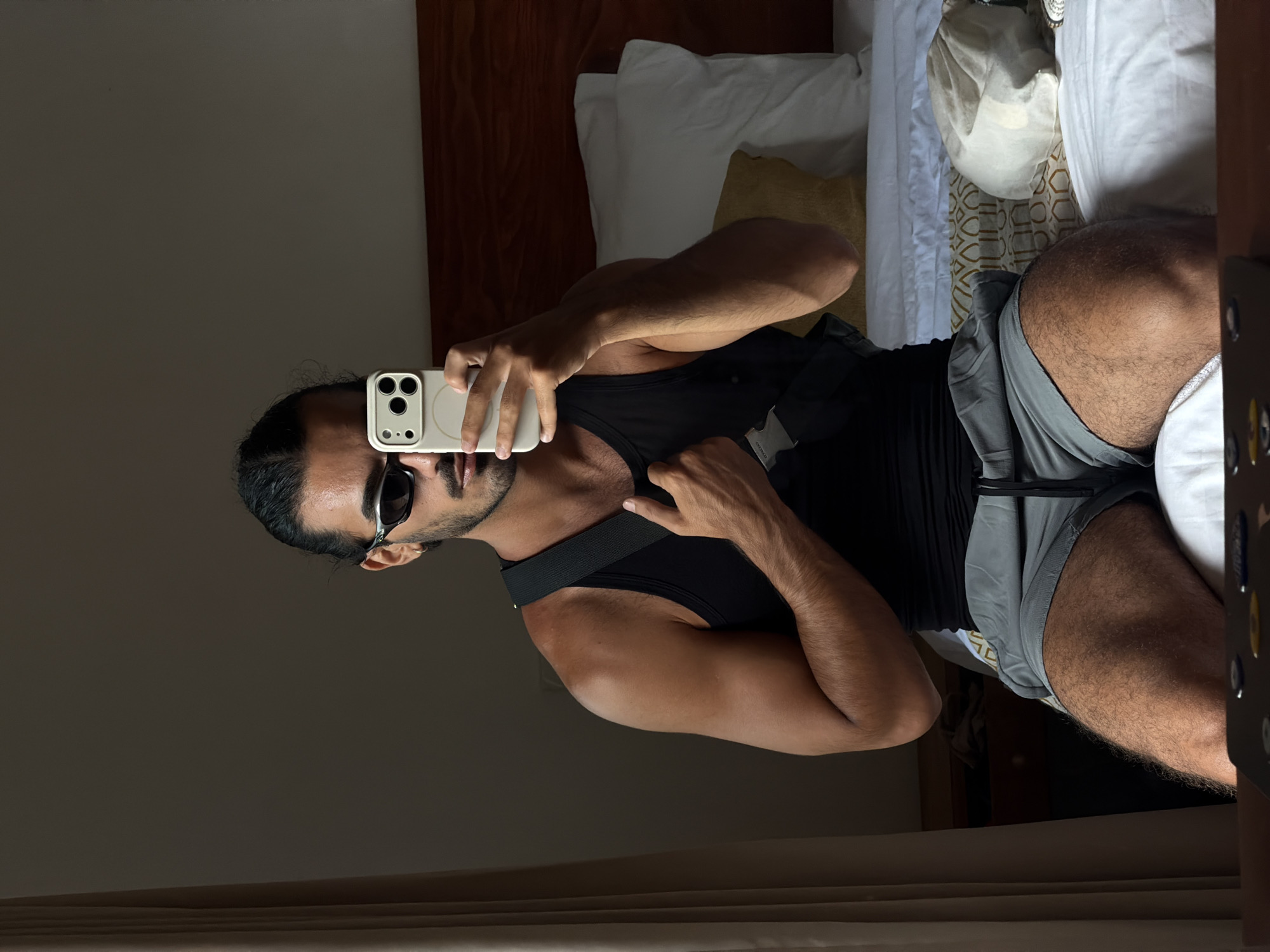 A man is captured in a mirror selfie inside what appears to be a bedroom or hotel room in North Kuta, Indonesia. He is holding a light-colored smartphone with a prominent camera array, taking the photo of his reflection. He has dark hair, styled back in a bun or ponytail, a beard or stubble, and is wearing dark sunglasses. His attire consists of a black sleeveless tank top and grey shorts. A black strap, possibly from a bag or part of his clothing, crosses his upper body. His arms are visibly muscular and tanned.
The background reveals a bed with white sheets and pillows, suggesting a comfortable sleeping area. A dark wooden headboard is partially visible behind the bed. Underneath the pillows, a patterned fabric, featuring a light background with a gold or brown geometric design, adds a decorative touch. A white hat or soft bag is resting on the bed to the right. The indoor lighting appears somewhat dim, creating a relaxed ambiance. No visible text is discernible in the image.