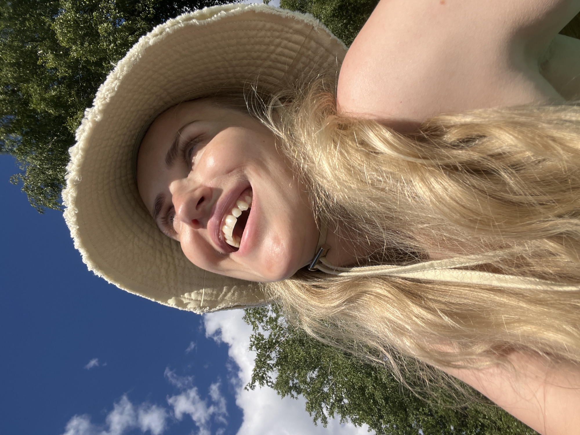 This is a close-up, angled, selfie-style shot of a young woman outdoors. She is wearing a light-colored, wide-brimmed hat, and her long, blonde hair is flowing. Her mouth is open in a wide smile, showing her teeth, and she is looking upwards and to her left. Sunlight illuminates her face, highlighting her features and casting shadows. The background consists of a bright blue sky with some scattered white clouds, and the green foliage of trees. The scene suggests a sunny day, possibly during the warmer months. The woman appears happy and relaxed. The overall impression is one of carefree enjoyment in an outdoor setting. The image does not contain any visible text. The location context of Saint Petersburg, Russia, combined with the sunny sky and foliage, suggests an outdoor activity or leisure time in a park or natural area.