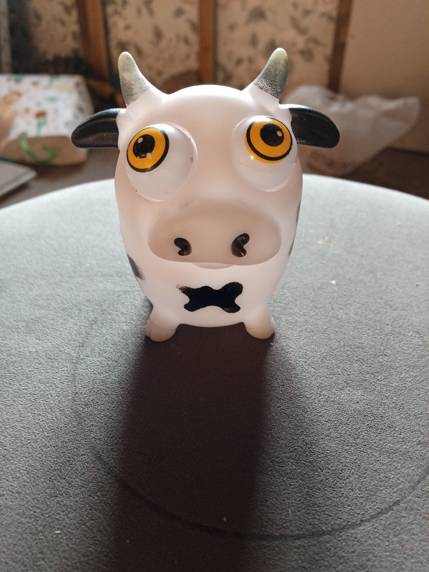 A white plastic toy cow with large yellow eyes and black spots sits on a textured gray surface. The cow has small black hooves, black ears, and two small horns. The background is blurred, showing what appears to be a room with furniture and possibly some fabric items. There are no people visible in the image, and no specific actions or interactions are depicted. The location is not identifiable as Krasny Yar, Russia.
