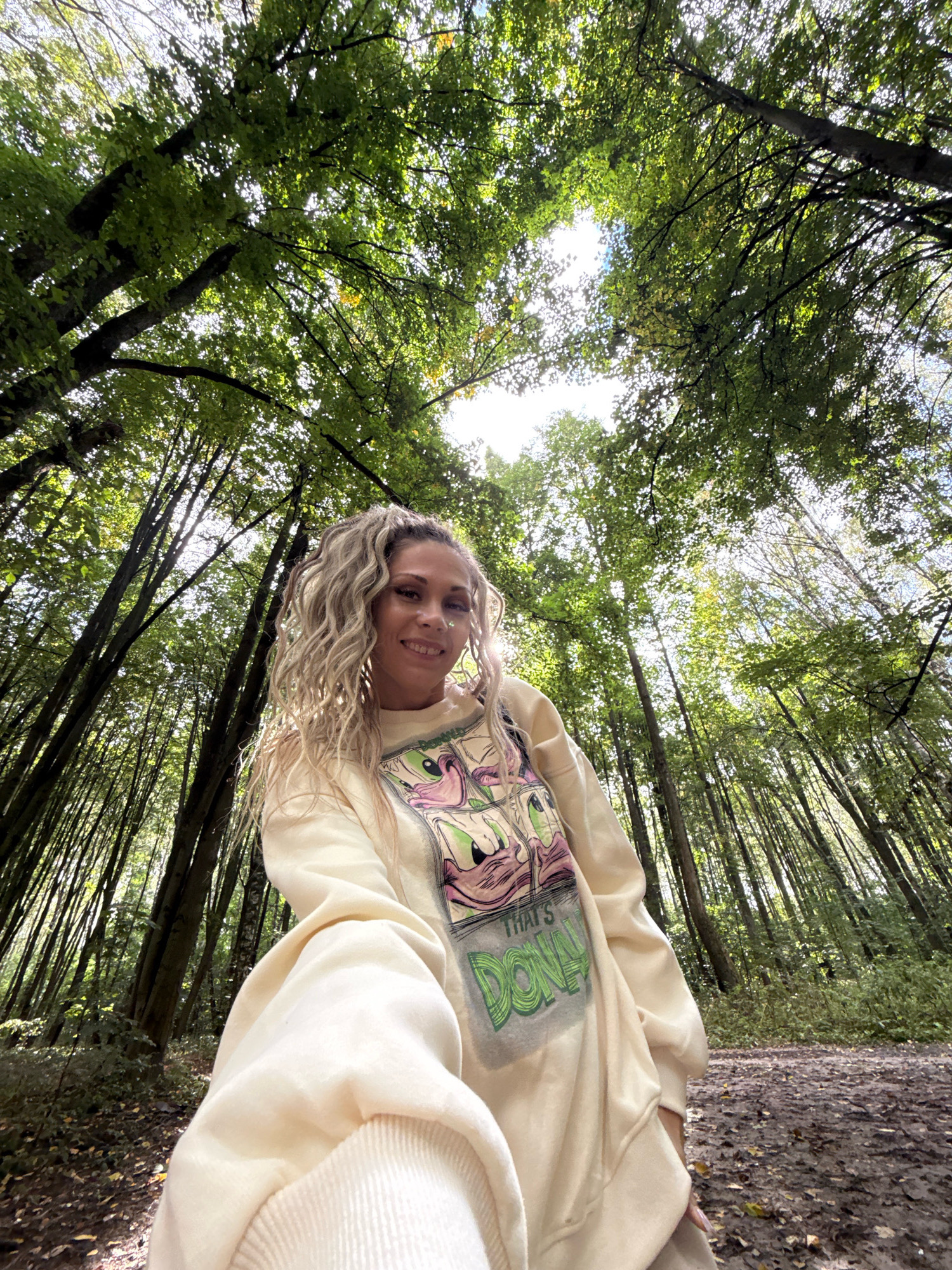 The image shows a woman standing in a forest. She has blonde, curly hair styled in a ponytail and is wearing a cream-colored sweatshirt with a cartoon graphic. The woman is smiling at the camera, which is positioned low, looking up at her and the canopy of trees. The trees are tall and densely packed, with green leaves forming a canopy overhead. Sunlight filters through the leaves, creating dappled light on the forest floor, which is covered in dirt and fallen leaves. The overall setting appears to be a natural, wooded area, possibly a park or trail. The location is specified as Rusanovo, Russia.