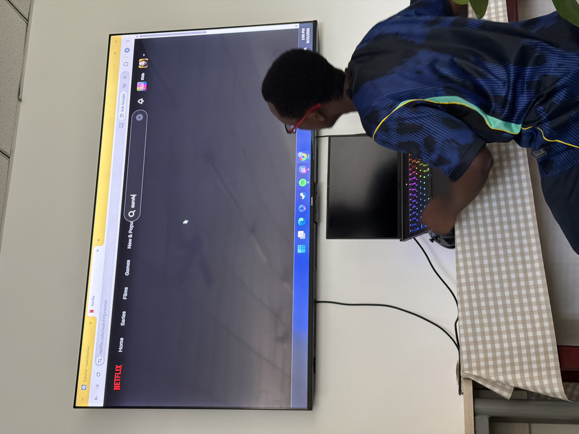 A young Black male wearing glasses and a blue and black patterned t-shirt sits at a table covered with a beige and white checkered tablecloth. He is facing away from the camera, looking at a large screen television mounted on a white wall. The television displays a Netflix search page with the search query "surviv" typed into the search bar. A laptop with a colorful illuminated keyboard is also on the table in front of the television. The room appears to be an indoor space, possibly an office or a home. The time on the television screen indicates 1:06 PM on 8/15/2026. The location is identified as Jiangjin, China.