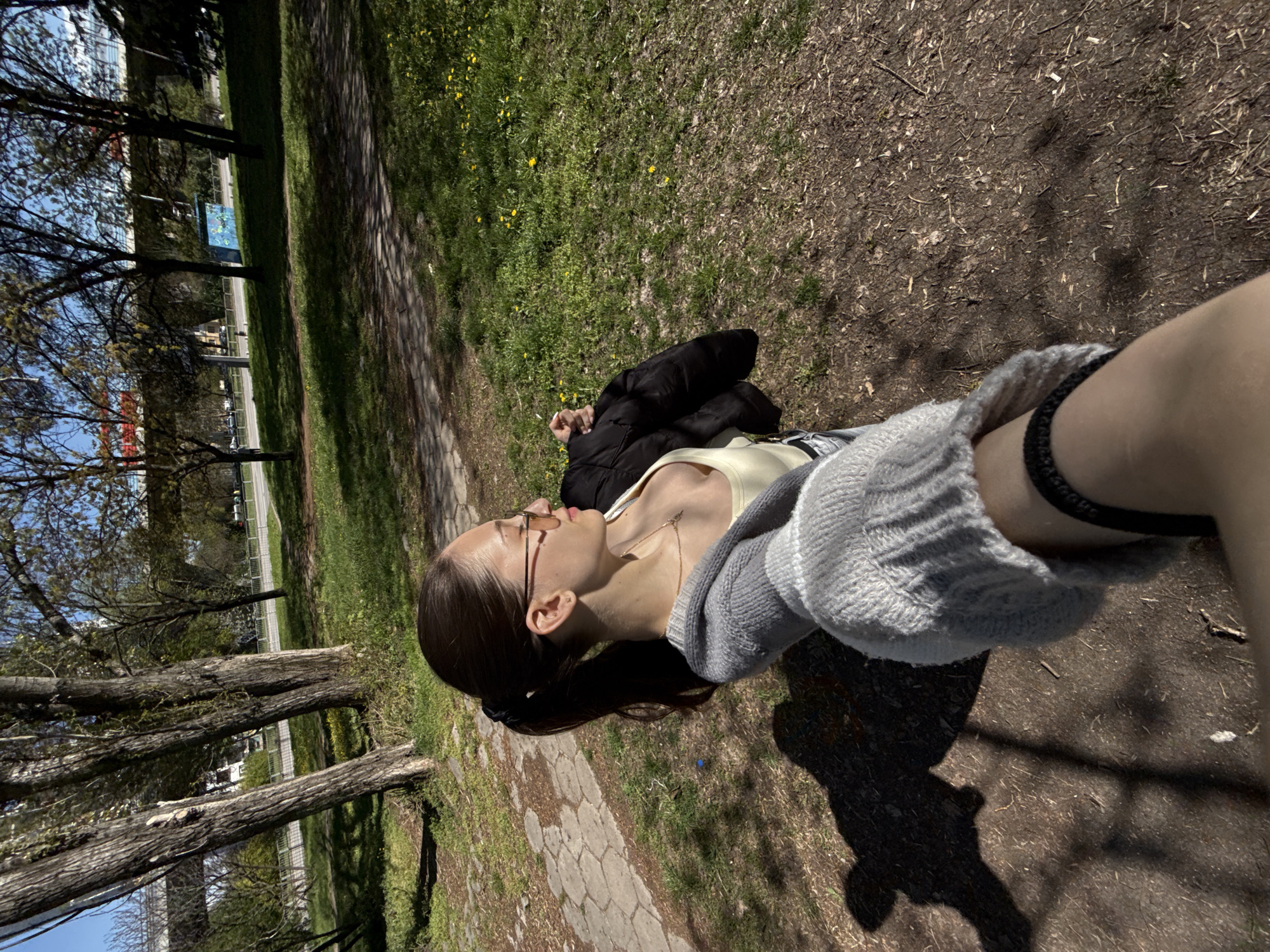 A vertical outdoor shot depicts a young woman lying on the grass in a park in Sofia, Bulgaria. Her head is towards the left, and her feet, which are partially visible, are towards the right. She is wearing glasses, a light-colored shirt, and a gray sweater. A gold necklace is visible around her neck. A black jacket is lying near her torso. The park has grass and trees. A road with vehicles and buildings can be seen in the background.