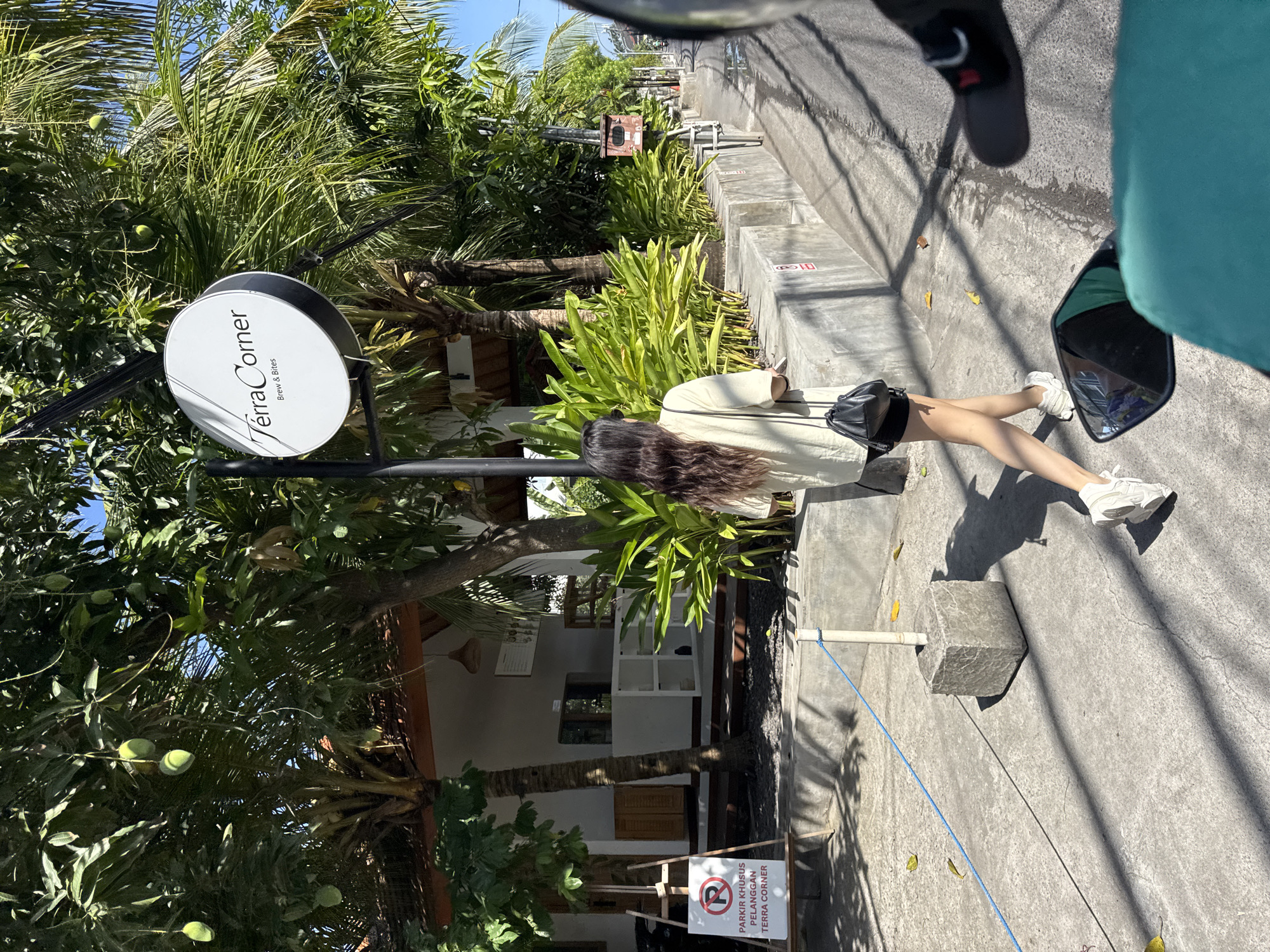 The photograph captures a street scene in Kuta, Indonesia, marked by a mix of tropical vegetation and urban elements.
In the foreground, a woman with long, dark hair is seen walking along the pavement. She is dressed in a light-colored top, a dark skirt, and white sneakers. Her shadow is cast onto the concrete sidewalk.
To the left of the woman, a sign for "Terra Corner Brew & Bites" is mounted on a pole, partially obscured by lush green trees laden with fruit. Further back, there is a building with light-colored walls and a wooden structure. A sign indicating reserved parking for "Terra Corner" customers is visible near the building.
The right side of the image reveals more of the paved street, concrete steps, and a curb. The view is partially obstructed by what appears to be parts of a motorcycle, including the mirror and a portion of the seat.
The overall scene is illuminated by bright sunlight, casting strong shadows and highlighting the tropical environment and urban setting.