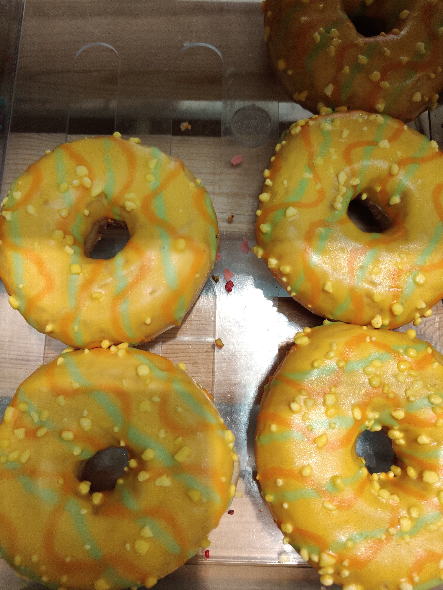 The image displays a close-up, overhead view of several brightly colored doughnuts arranged in a clear display case. The doughnuts are the primary subjects, with at least five visible, filling most of the frame.
Each doughnut features a vibrant yellow icing base, adorned with swirling, irregular stripes of green and orange. They are generously topped with small, irregular, yellowish sprinkles, which appear to be crunchy or candy-like. All doughnuts have a classic ring shape with a central hole. The overall color scheme is lively and tropical.
The doughnuts are presented in a transparent container or display case, through which a light brown, possibly wooden, surface is visible underneath. Some small, scattered specks of pink, white, and red debris are visible on the surface of the display case, suggesting a retail environment such as a bakery or cafe. The lighting appears to be artificial and illuminates the doughnuts evenly.
No people are visible in the image. The activity depicted is the display of these confectionery items, likely for sale. There is no visible text. The time of day and weather cannot be determined from the image.