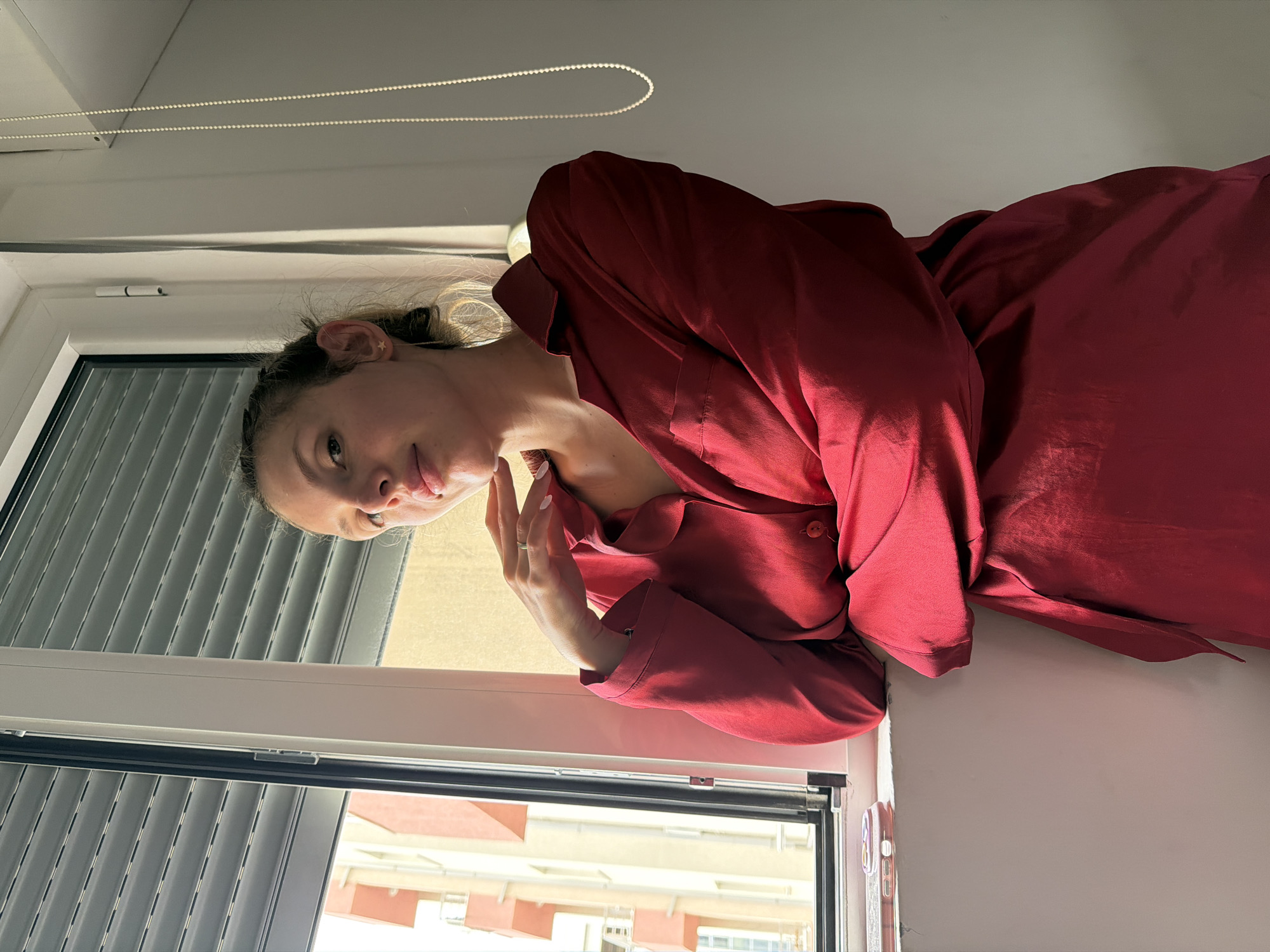 A young woman wearing a red dress stands next to a window with the blinds partially open, gazing into the camera. Outside the window, a multi-story, light-colored building with red accents can be seen. Based on architectural styles, color palettes, and the prevalence of certain building materials in the region, the location could be in Budva, Montenegro.