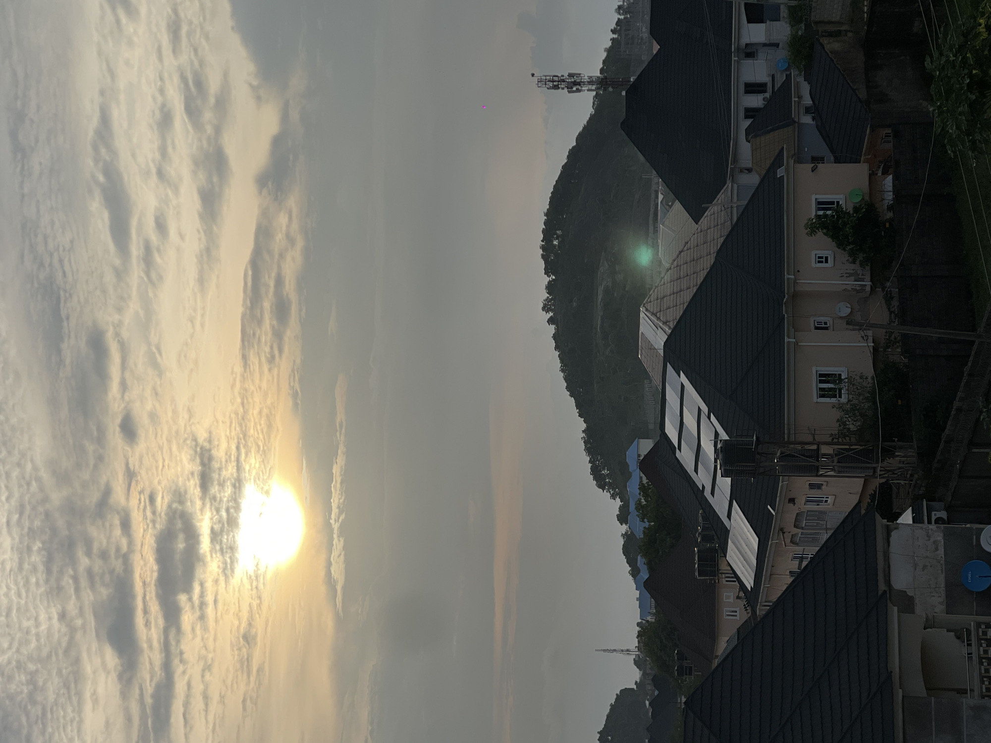 The image captures a view of residential buildings in an urban area, likely a suburb or a developing district, under a cloudy sky with the sun peeking through. The scene appears to be an aerial or elevated perspective, looking over a collection of houses with dark, sloped roofs, many of which are tiled. Some houses have solar panels installed on their roofs, indicating modern infrastructure. In the background, a lush green hill or mountain range rises, with trees covering its slopes. A communication tower stands prominently on the right side of the hill, and a smaller pink object is visible in the sky to the left of the tower. The lighting suggests either dawn or dusk, with the sun casting a soft glow through the clouds. The overall impression is one of a quiet, residential neighborhood in a city characterized by a mix of modern housing and natural landscape. This scenery is consistent with descriptions of many developing areas and suburbs within the city of Abuja, Nigeria, known for its planned urban layout and surrounding natural topography.