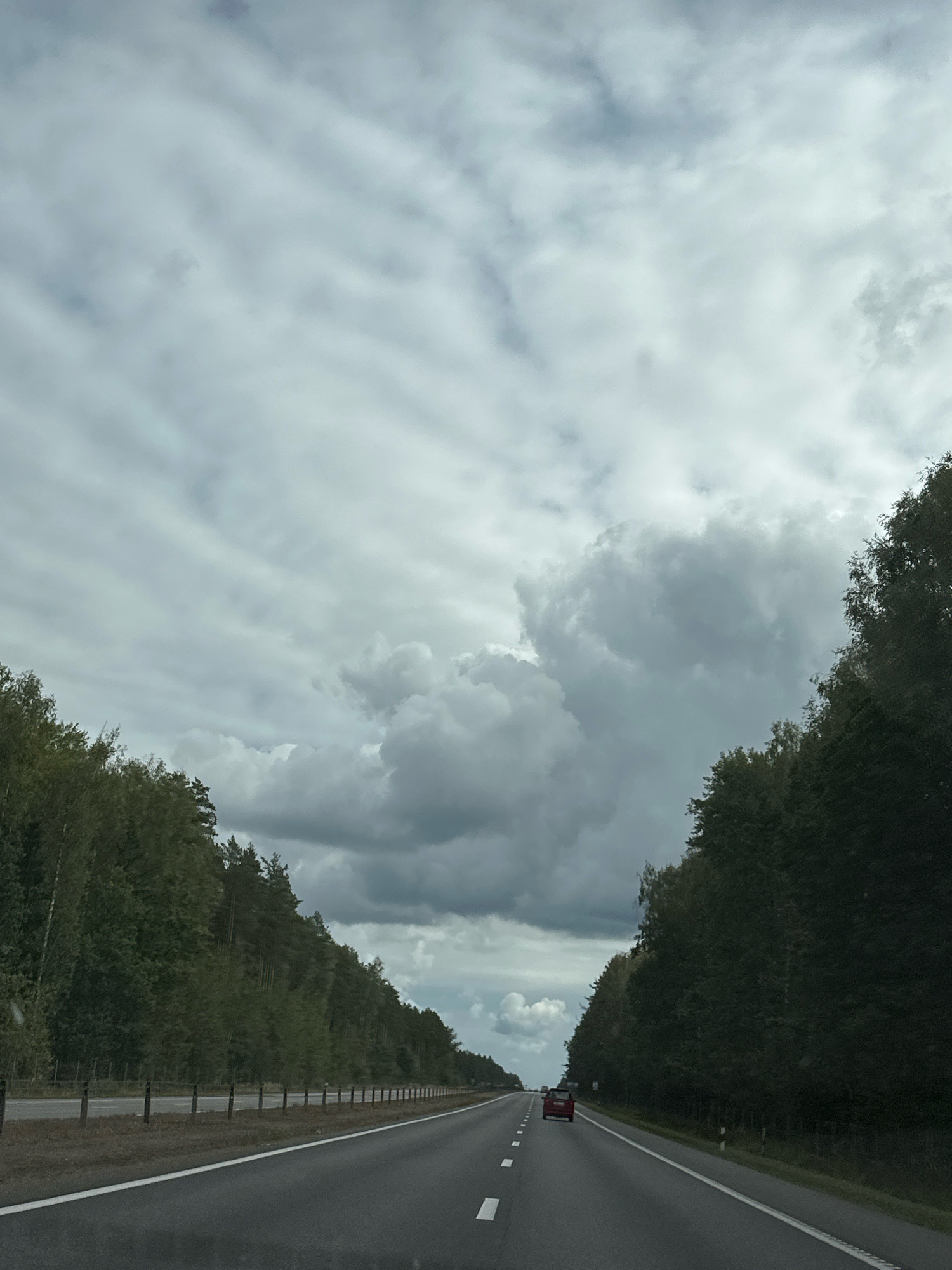 The image shows a view from inside a vehicle traveling on a highway through a rural area. The highway is asphalt with white lane markings. On either side of the highway are dense forests with a mix of deciduous and coniferous trees. The sky is overcast with heavy, gray clouds, suggesting impending rain or a stormy day. A red car is visible in the distance on the highway, traveling in the same direction. The scene appears to be located in the Вяззеўскі сельскі Савет, Belarus, characterized by its extensive forests and typical rural Belarusian highway infrastructure, including roadside markers and a simple fence line along the left side of the road.