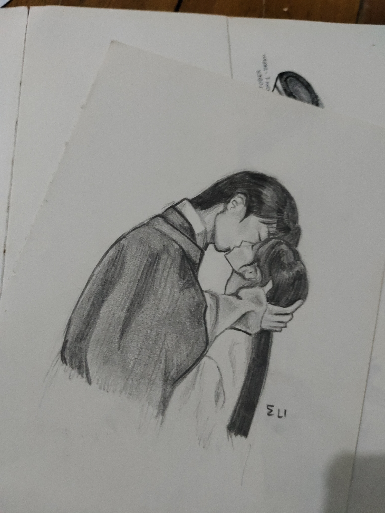 The image displays a slightly angled, overhead photograph of a pencil drawing on a sheet of white paper. The drawing depicts two human figures in an embrace, engaged in a kiss. The figure on the left, appearing to be male, has short, dark hair, wears glasses, and is dressed in a dark, long-sleeved garment with a collar. He is leaning downwards towards the other figure. The figure on the right, appearing to be female, has long, dark hair flowing down her back and is wearing a lighter-colored garment. Her face is turned upwards, and her left hand is gently placed on the side of the male figure's head or jaw. Both figures are rendered in shades of grey using pencil, with distinct outlines and shading to indicate depth and form. In the bottom right corner of the drawing, the letters "ELI" are written vertically.
The sheet with the drawing is positioned on a light brown surface, likely a wooden table or desk. To the upper right of the main drawing, on a separate piece of paper, there is a small, faint pencil sketch resembling an ear, alongside some handwritten text, partially legible as "JOBER" and "DAY 1". Additional sheets of plain white paper are visible on the left and top edges of the frame. The overall scene is an indoor photographic record of an artistic creation. No specific city or geographic location is identifiable from the depicted drawing or its immediate photographic context.