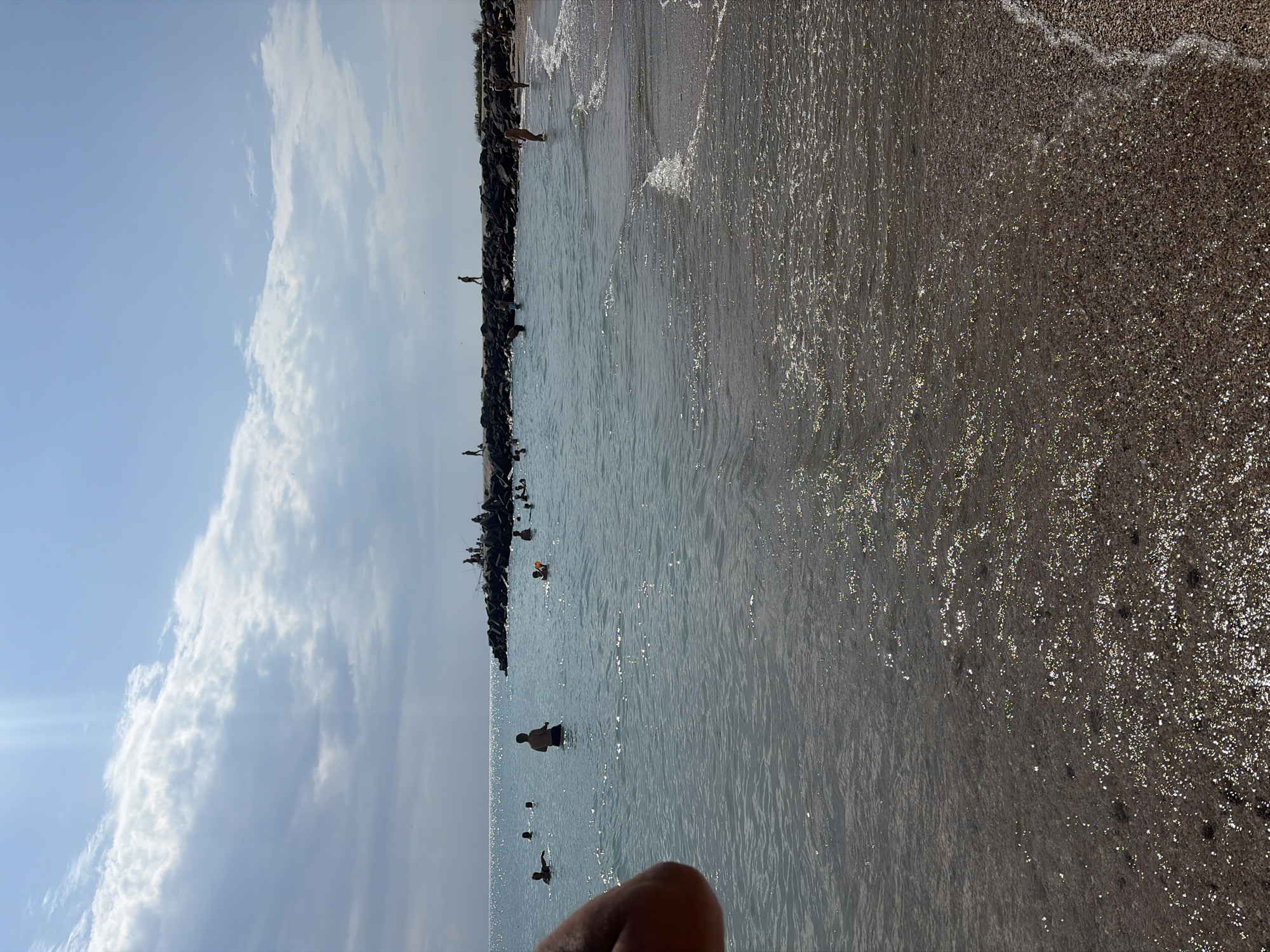 This image captures a sunny day at a beach in Pomorie, Bulgaria. A group of people are seen swimming and wading in the clear, shallow waters of the Black Sea. Some individuals are standing along a breakwater made of large rocks, where a few are fishing. The beach itself is sandy and appears to be lightly populated. The sky is bright blue with scattered white clouds, and sunlight glints off the water's surface. The overall scene depicts a leisurely day at the seaside.