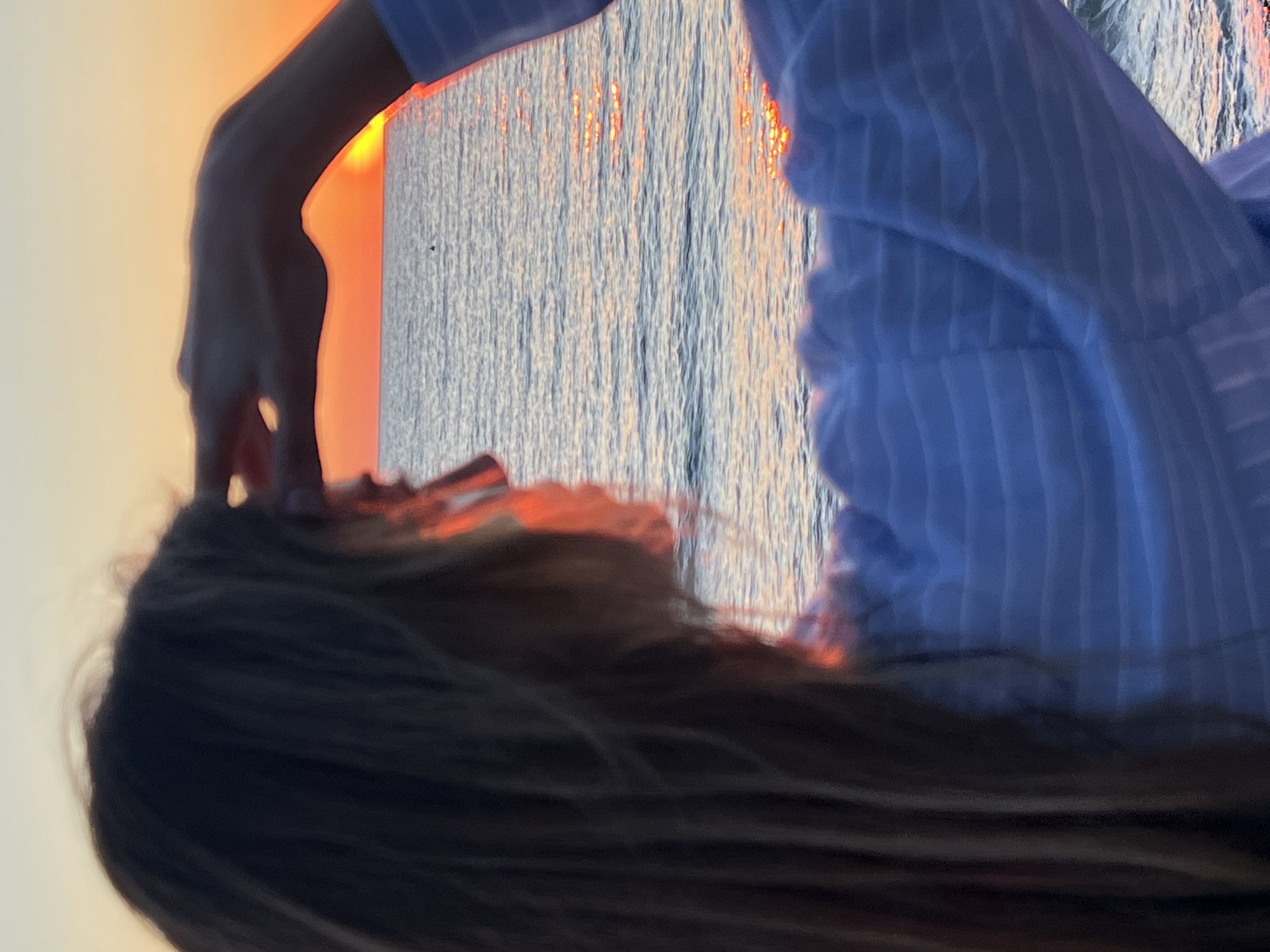 A waist-up, eye-level, portrait photograph shows a woman with her back to the camera, facing the Adriatic Sea during sunset, taken in the city of Bar, Montenegro. Her blonde hair falls downwards, partly obscuring her shoulders, face and the horizon. Her right arm is raised, with the hand positioned near the top of her head. She wears a horizontally striped blue shirt. The sea is visible in the background, with the reflection of the setting sun creating streaks of orange light on the water's surface. The sky is orange with an orange gradient.