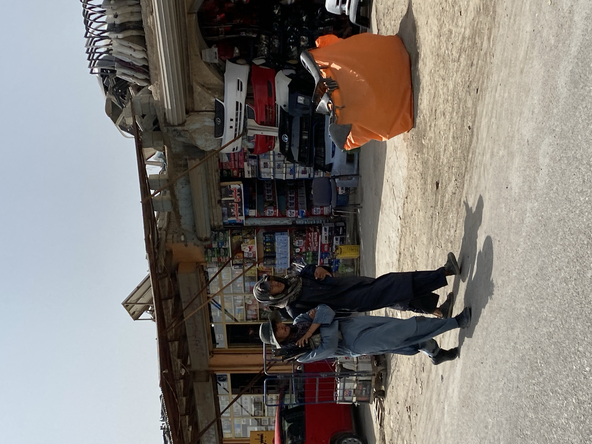 The image depicts two individuals walking on a dirt road in front of a storefront in the city of Kandahar, Afghanistan. The storefront appears to be a shop selling car parts, with various car bumpers stacked on shelves and leaning against the building. In the foreground, the two individuals are walking from left to right. The person on the left is wearing a light blue traditional shalwar kameez and a wide-brimmed hat, and is carrying a bundle of what appear to be long, slender objects. The person on the right is wearing a dark, hooded garment. Behind them, the street is lined with shops and displays of goods. An orange tarp-covered object is visible on the right side of the image, in front of the shop. The sky is clear and blue, suggesting a sunny day. The overall environment appears to be a bustling commercial area, potentially a market street.