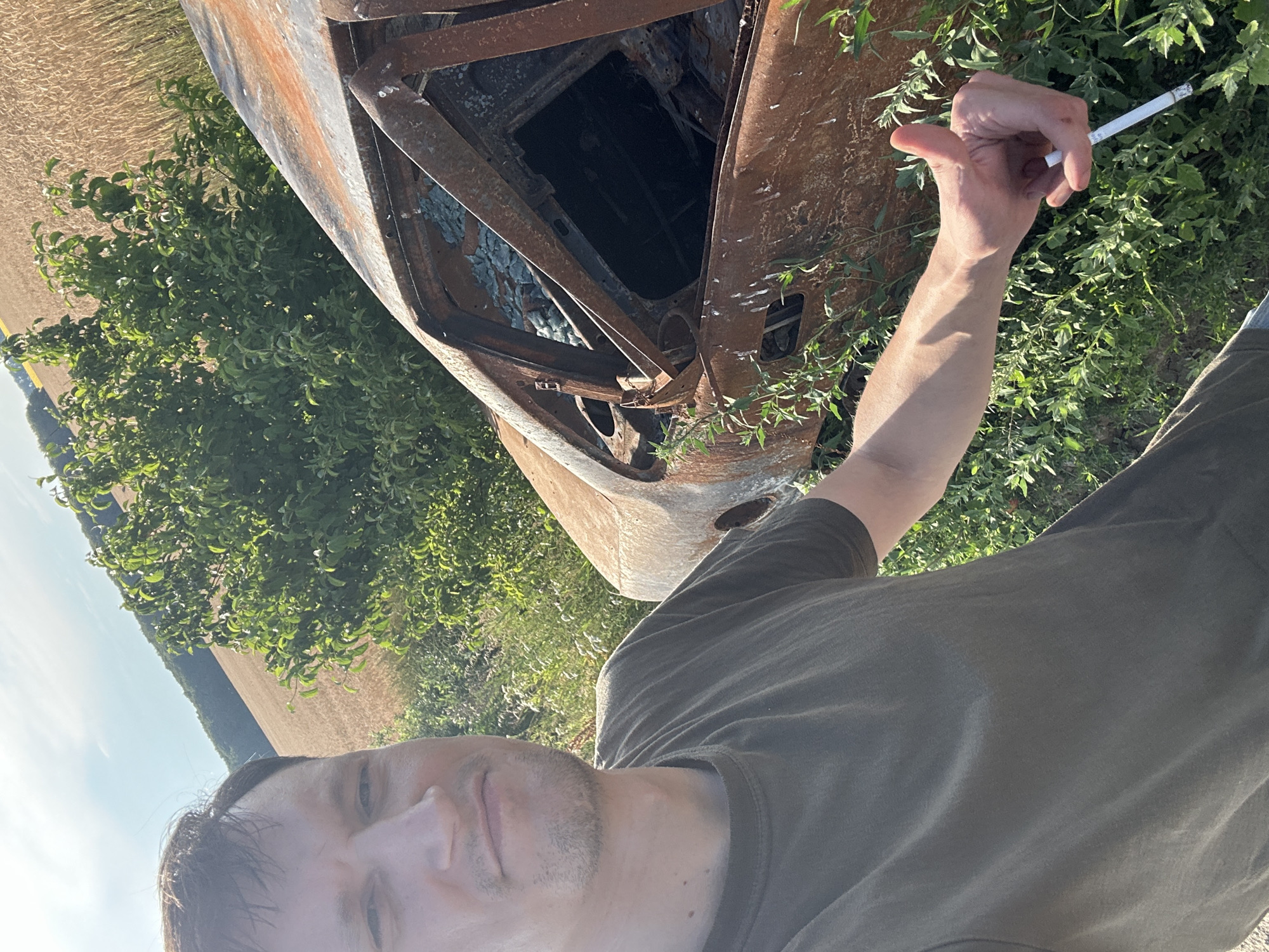 A selfie taken by a Caucasian man in his 30s shows him giving a thumbs-up next to a burnt-out car in Makiv, Ukraine. The man is wearing a dark olive t-shirt and has short brown hair. The car, which appears to be a sedan, is heavily damaged and rusted, with its windows shattered. The scene is set in a rural area with a field of wheat or similar grain in the background, bordered by a line of trees and a clear blue sky. The foreground features green vegetation growing around the car. The man's left hand is visible, holding a cigarette.