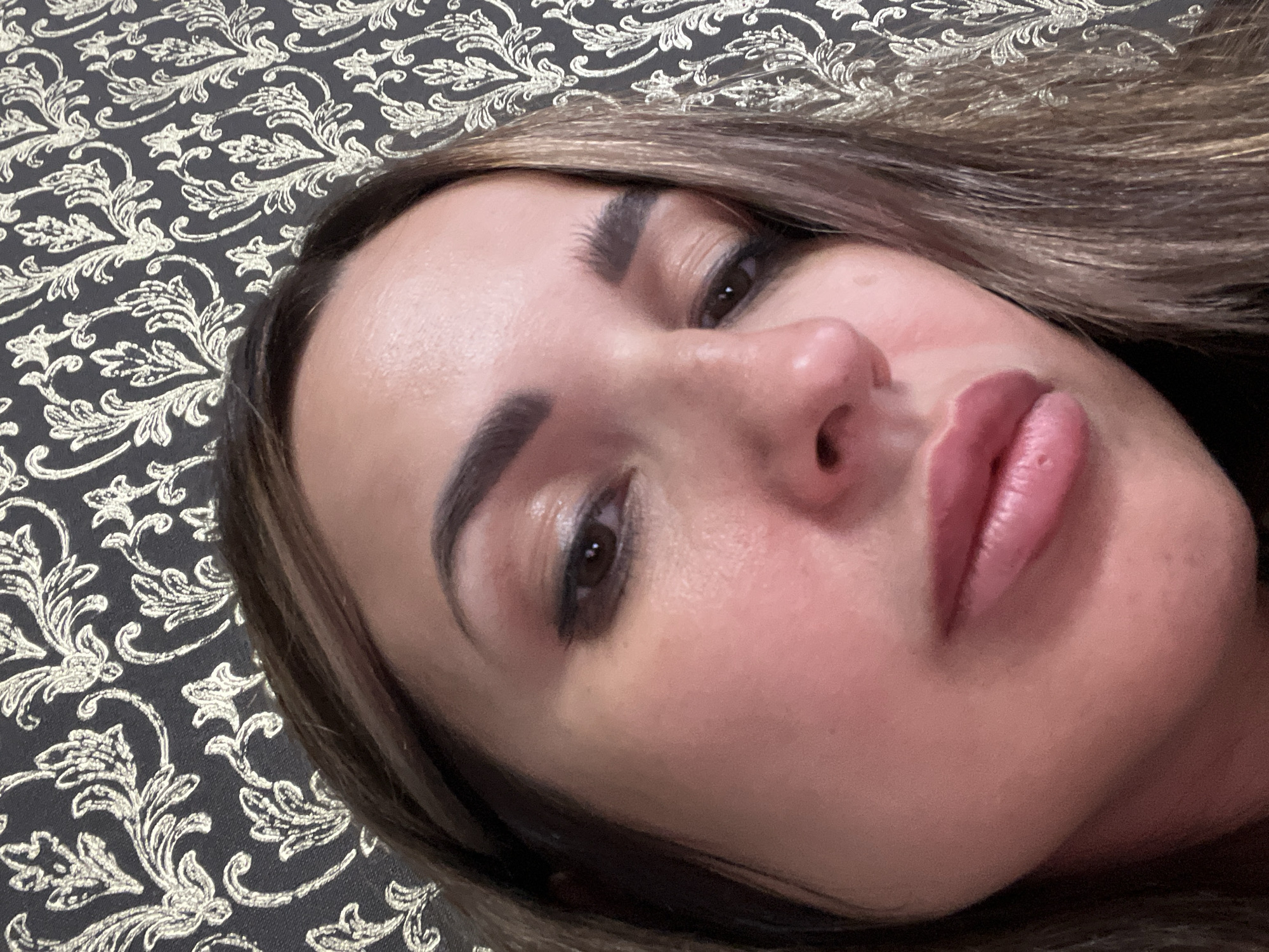 A close-up, upward-angled portrait shows the face of a woman with brown hair against a dark background with a gold floral pattern. She is wearing makeup, including eyeshadow and lipstick. The lighting is soft, highlighting her facial features and skin texture. The image does not provide information about a specific city or location.