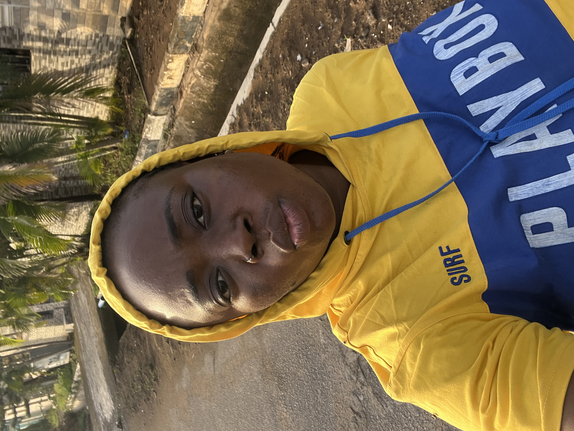 A young Black person wearing a yellow and blue hooded sweatshirt with the words "PLAY BOY" and "SURF" printed on it is posing for a selfie outdoors. They are wearing the hood up. The background includes a street, a building with a tiled wall, and some trees, possibly palm trees. The location is likely somewhere in Abuja, Nigeria, given the prompt's specification.