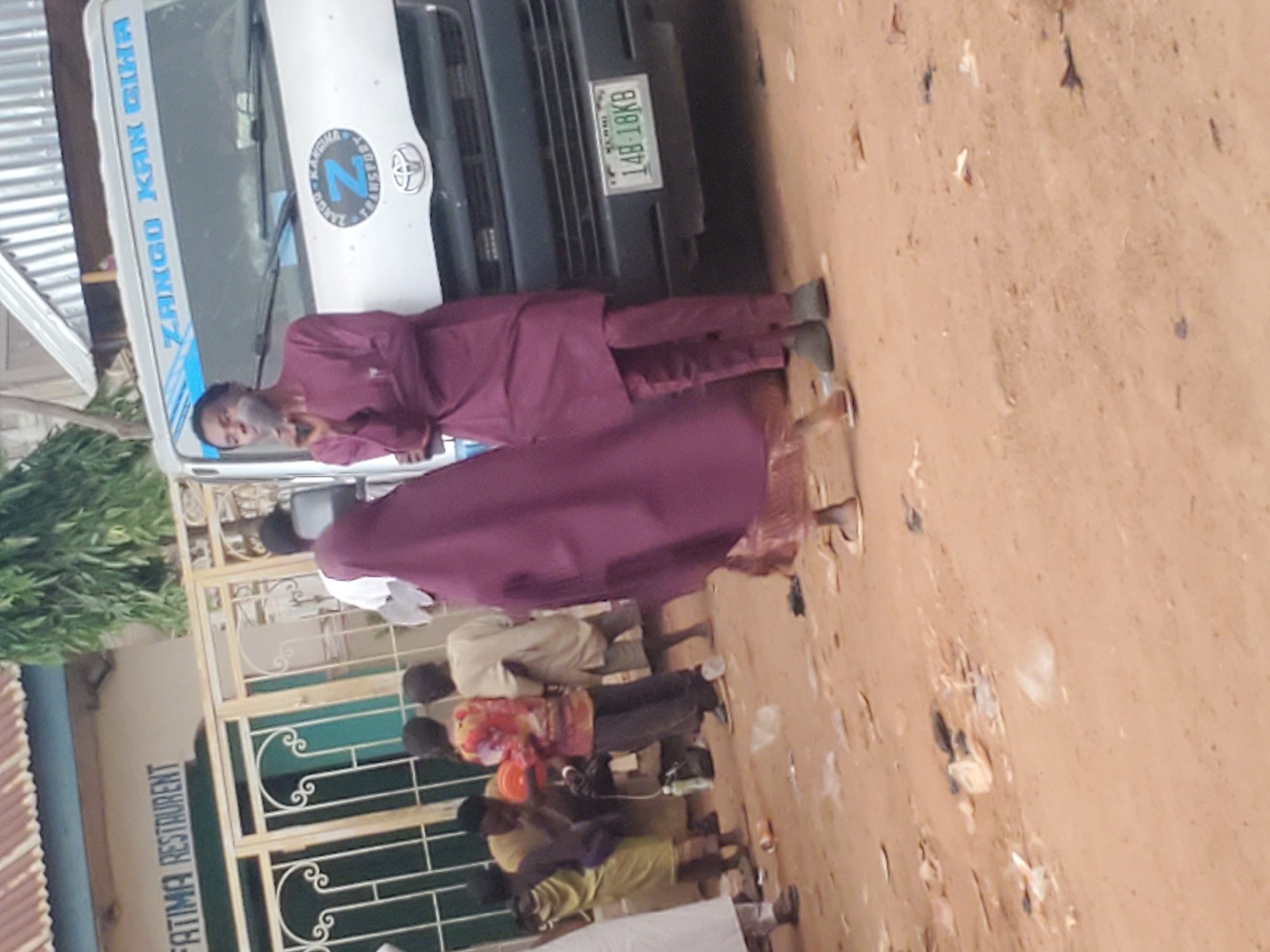 The image depicts an outdoor scene in Funtua, Nigeria, likely in front of a business. A white van with blue lettering that reads "ZANGO KAN GIHA" and a circular emblem with a "Z" is parked on a dirt or gravel surface. License plate "14B-18KB" is visible on the front of the van. To the left of the van, a building with a corrugated metal roof and a sign that partially reads "FATIMA RESTAURANT" is visible. A decorative metal gate is in front of the restaurant.
Several individuals are present in the scene. In the foreground, a person in a maroon outfit, including a long tunic and pants, stands facing towards the van, holding what appears to be a cigarette. Another person in a similar maroon garment is partially visible to the left, also appearing to be in motion. Further back, several other people are gathered. One individual in a colorful patterned shirt and dark pants is holding a red spherical object. Another person in a light-colored tunic and shorts stands nearby. A person in a yellow outfit is visible to the left, and a person in a white garment is partially seen in the far left. The ground is sandy and littered with debris, including plastic wrappers and discarded items. The overall impression is of a public space with moderate activity.