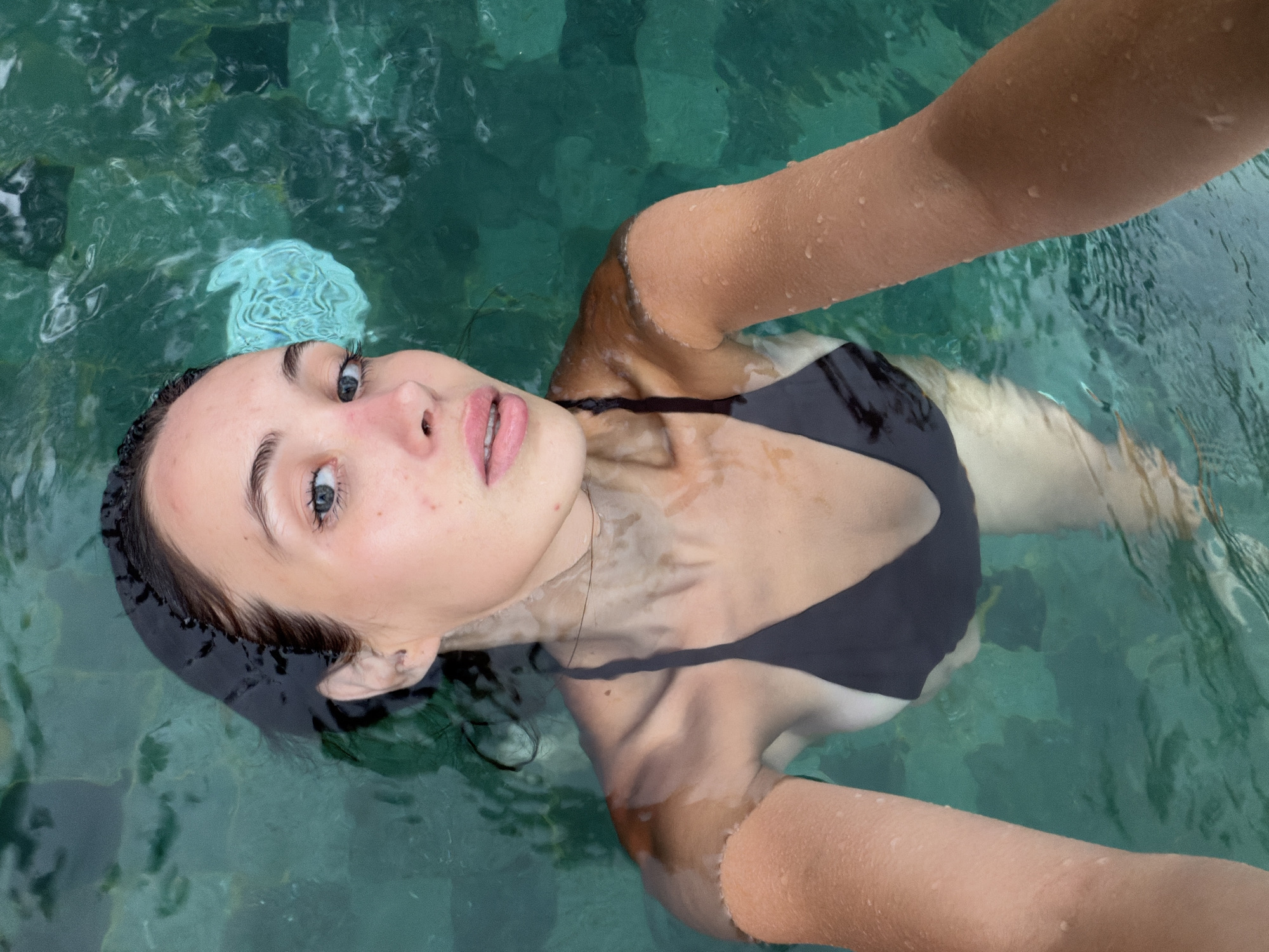 A woman is floating in a pool of water. She has a fair complexion, dark hair, and is wearing a black bikini top. Her arms are extended on either side of her body, resting on the surface of the water. The water is a greenish-blue color and appears clear. A turquoise-colored flower adorns the top of her head. The location is identified as the Ubud District, Indonesia, based on the pool design, water color, and the ethnicity of the woman. The image appears to be a selfie, with the woman holding the camera at arm's length above her head.