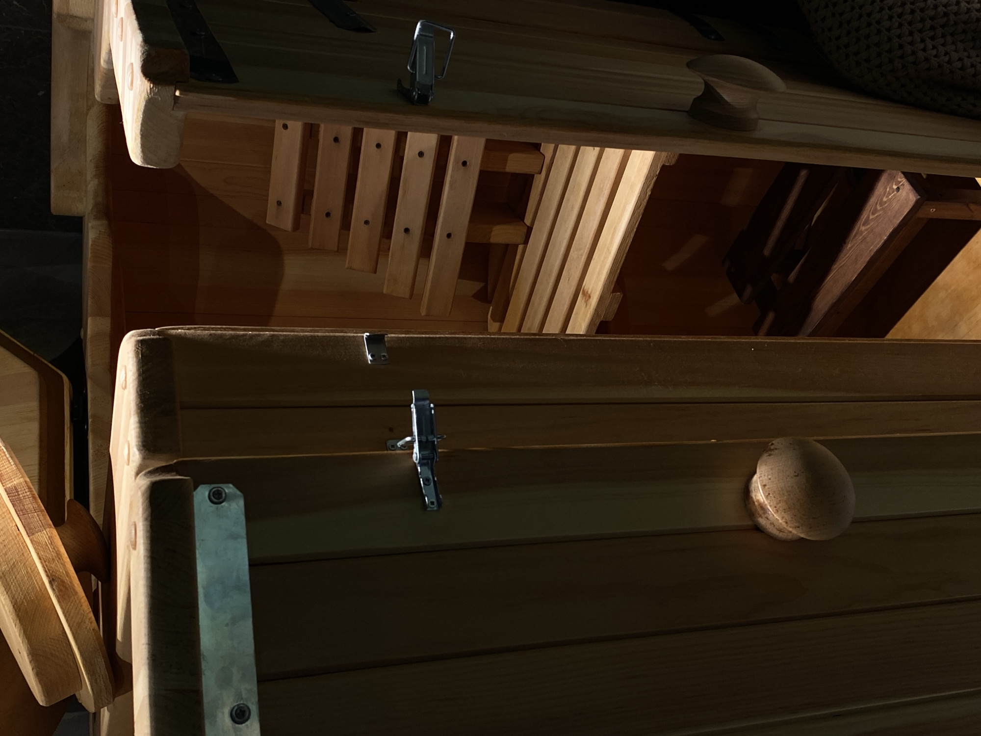 This image displays a close-up view of interior wooden structures, likely within a sauna or a traditional Russian banya, given the provided location context of Votkinsk, Russia. The composition features two main wooden elements, resembling benches or shelves, stacked vertically in the frame.
The upper wooden structure is equipped with two prominent metal latches, one on the left and another centrally located. To the right, a round, wooden knob is visible, partially obscured by a dark, textured, possibly woven or knitted material. Below this upper structure, a series of light-colored wooden slats or rungs are arranged, secured by small, dark screws, suggesting a slatted base, a ladder component, or part of a ventilation system.
The lower wooden structure also features a single metal latch on its upper left edge and a round, wooden knob on its right side. The entire scene is bathed in dim, dramatic lighting, casting strong shadows and highlighting the grain and smooth finish of the light-colored wood. The warm tones of the wood are contrasted by the cooler metallic sheen of the latches. No people or specific activities are taking place; the scene is static, showcasing the detailed craftsmanship of the wooden interior. There is no visible text in the image.