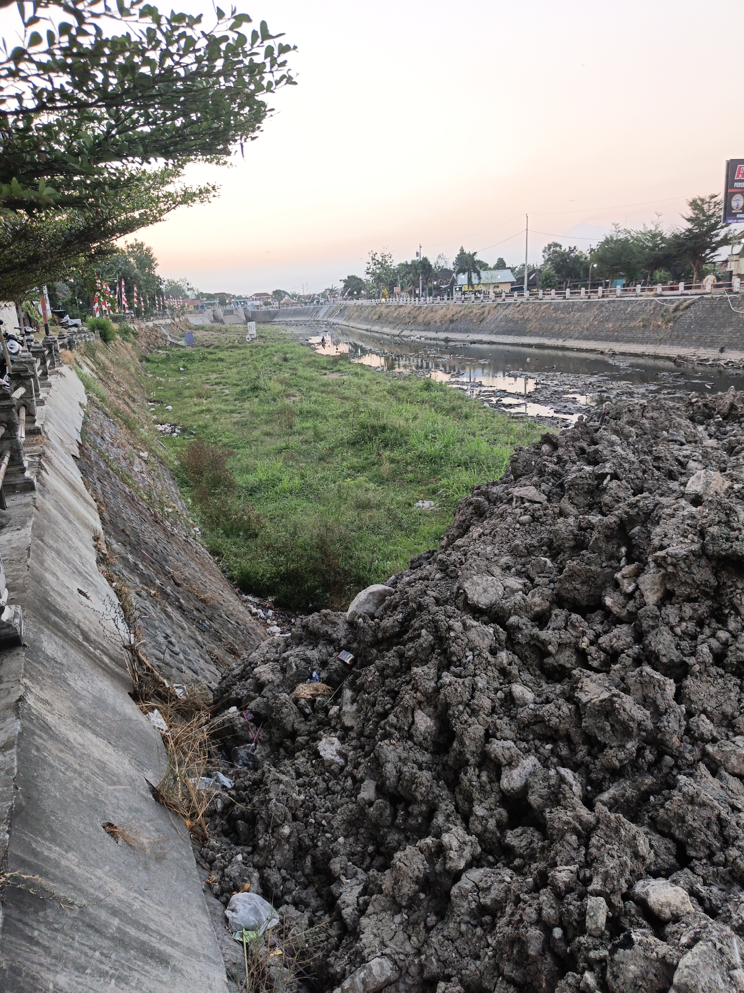 The image depicts a canal in the city of Planggu, Indonesia, during twilight. A large pile of dark soil or mud occupies the foreground, obscuring much of the canal's bank on the right. The canal itself is shallow, with a muddy bottom and a thin layer of water reflecting the soft hues of the sky. Patches of green grass and weeds grow along the canal's edge and in the dry areas. On the left side of the canal, a concrete retaining wall slopes down towards the water, with loose stones and dried vegetation visible along its base. Beyond the canal, a street is lined with buildings and trees, and several red and white flags are displayed, suggesting a public event or national celebration. A few motorcycles are parked near the flags. The sky transitions from a pale orange near the horizon to a lighter shade of pink and then white. In the distance, hints of mountains can be seen. There are no people prominently featured in the image, though a few indistinct figures might be present in the canal or on the far bank. The overall scene suggests an area undergoing maintenance or construction, or simply a natural landscape in an urban setting.